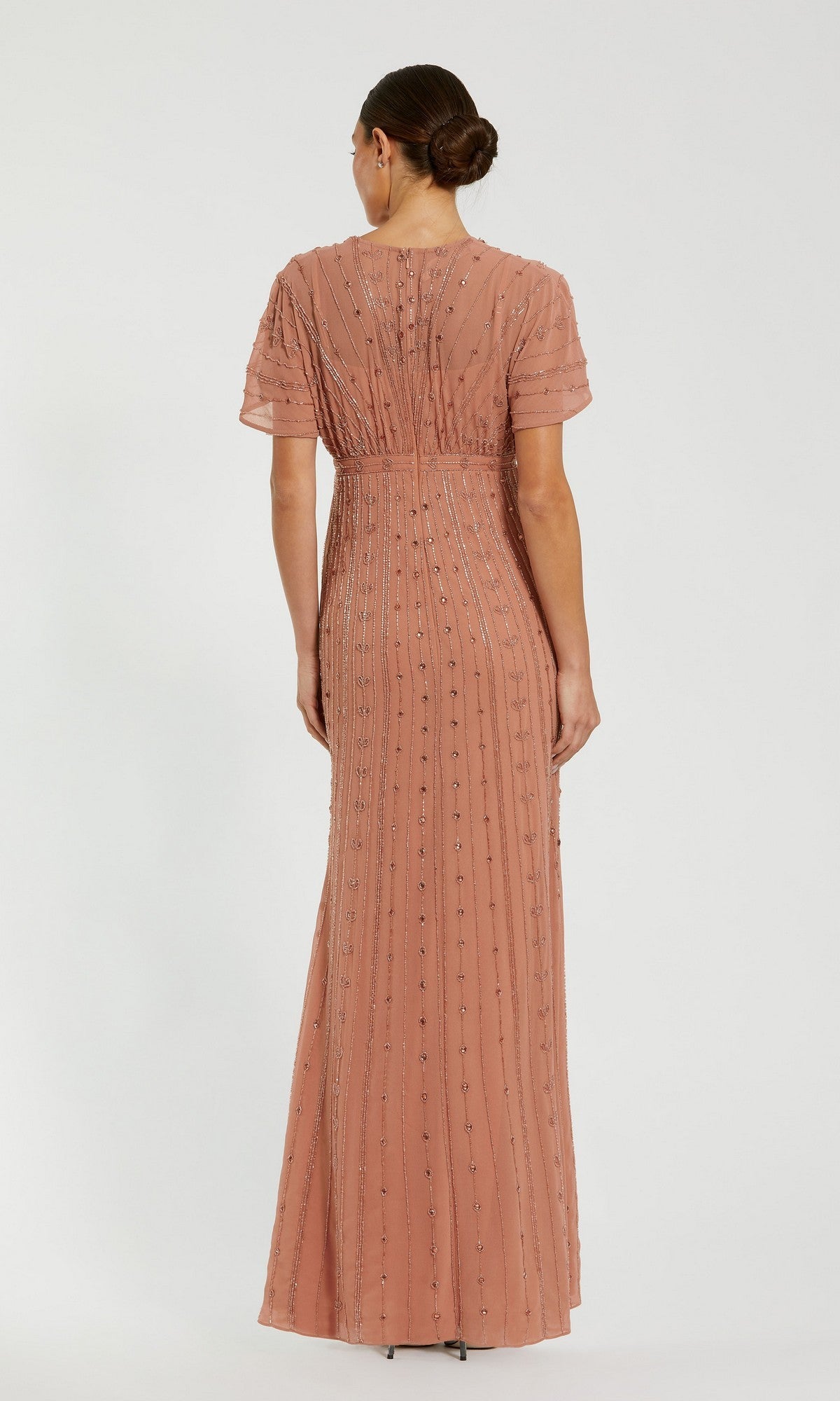 A woman stands with her back to the camera, wearing the Mac Duggal 94149 long formal dress—a floor-length gown in dusty pink featuring short sleeves and beaded vertical lines. Her hair is styled in a low bun, completing an elegant ensemble.