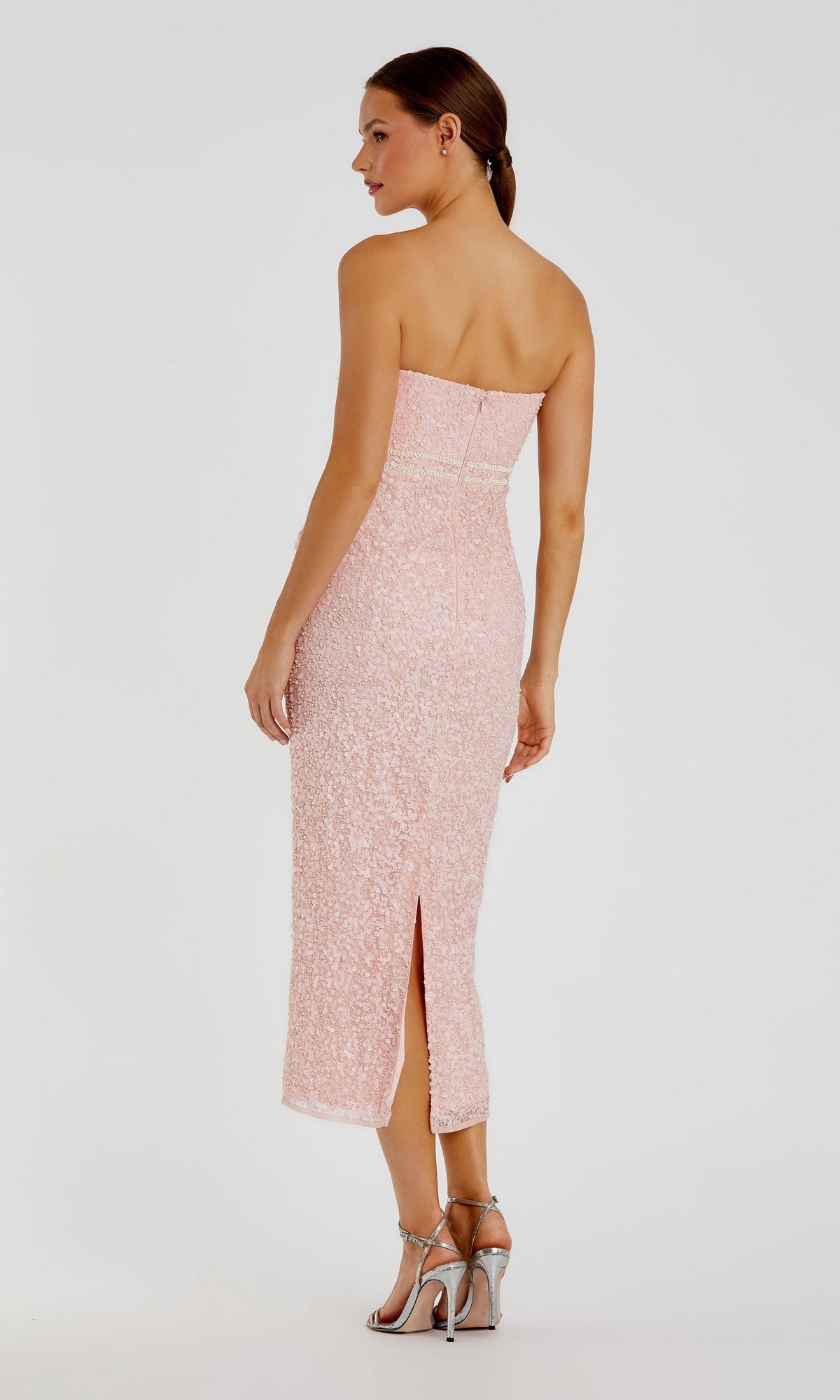 A woman wearing the Mac Duggal 94200 tea-length party dress—a strapless design adorned with light pink sequins reminiscent of a pink pearl-embellished gown—with a back slit and paired with silver high heels, stands against a plain white background.