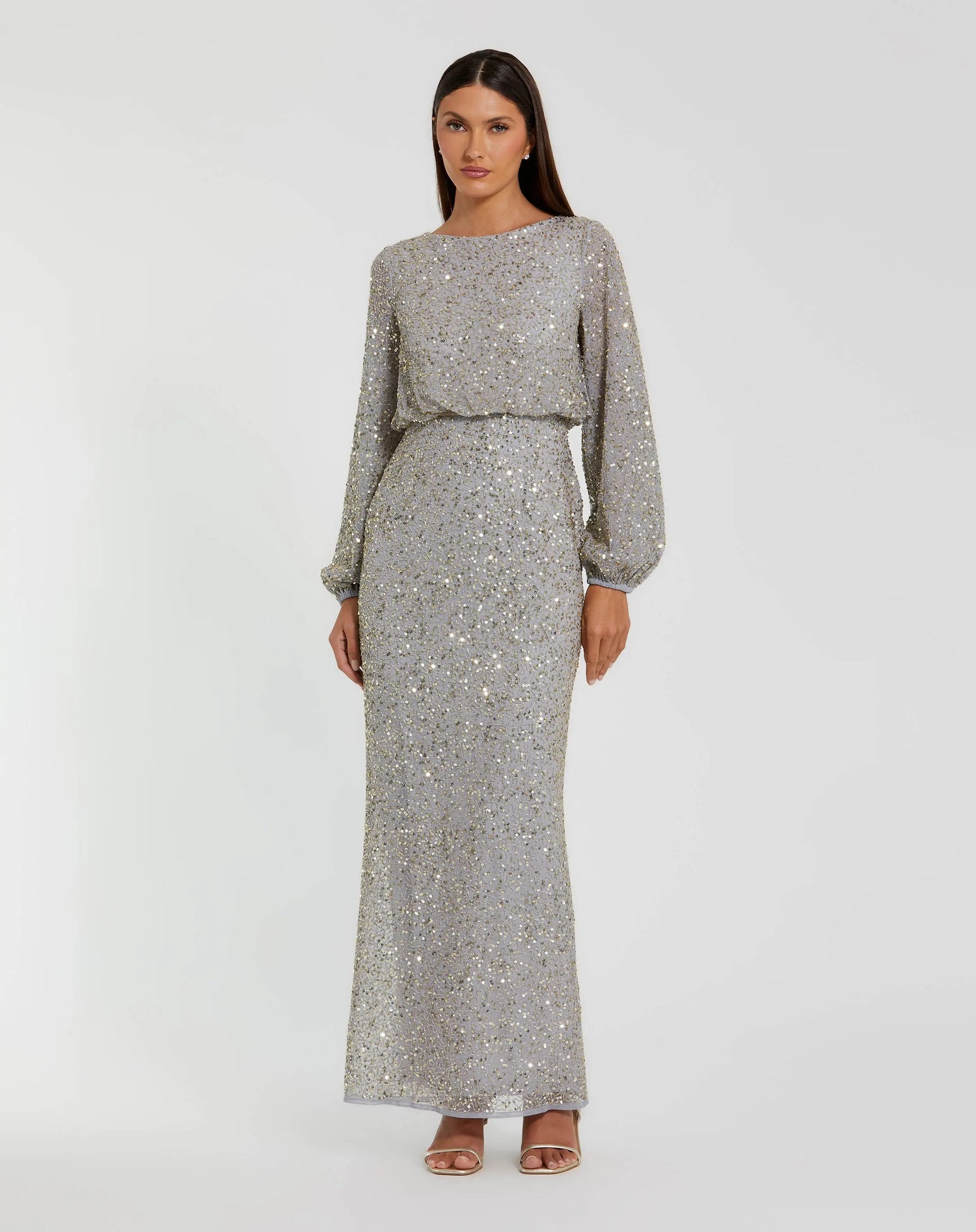 A woman stands in a full-length, long-sleeve platinum gold gown—Mac Duggal 94203—with a cinched waist and open-toe heels, set against a plain white background. This elegant sequin dress from Mac Duggal exudes both glamour and the sophistication of a modest long formal dress.
