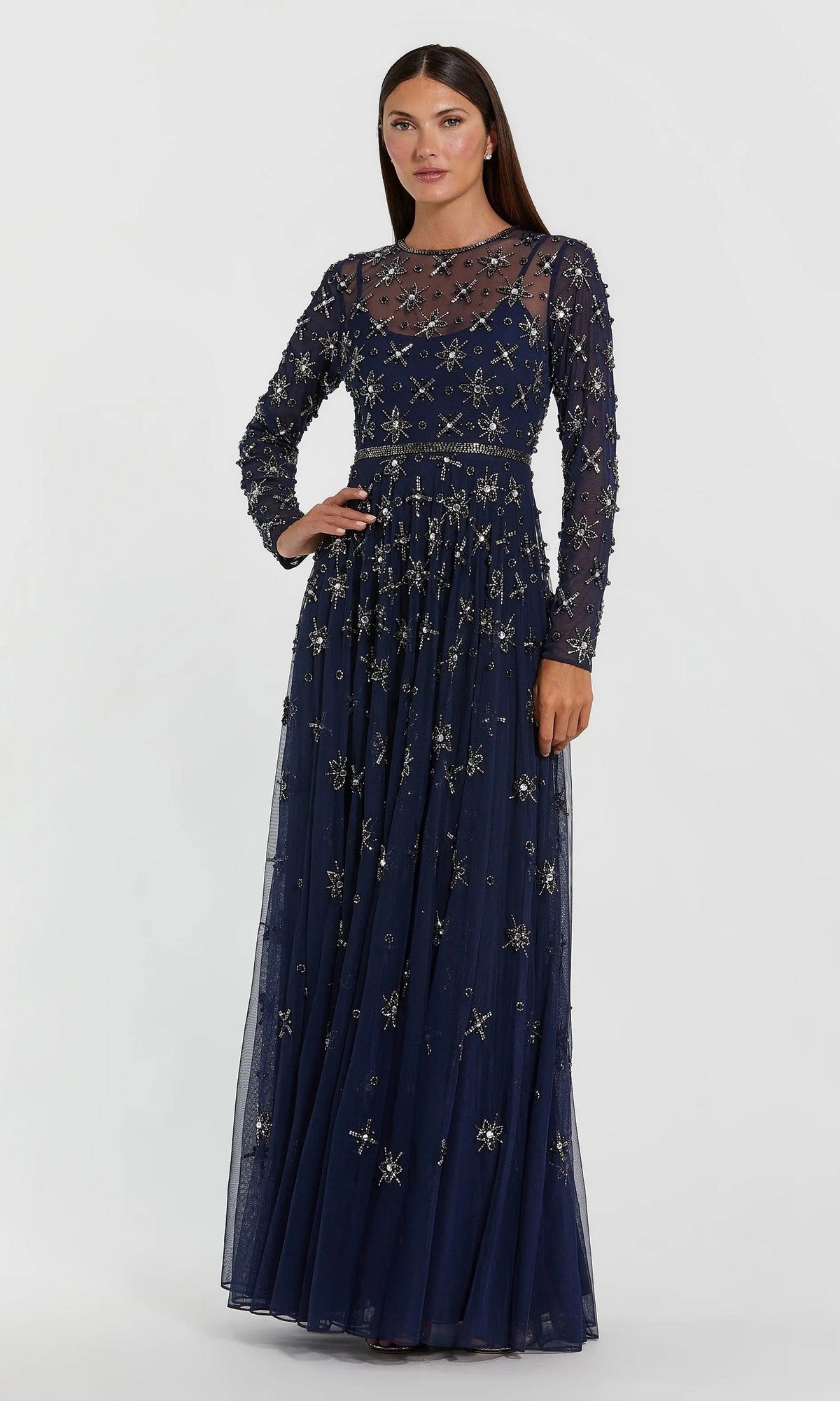 A woman wearing the Long Formal Dress: Mac Duggal 94216 in midnight—a floor-length gown featuring sheer long sleeves, intricate gold embroidery, and delicate beaded embellishments—poses against a plain, light background.