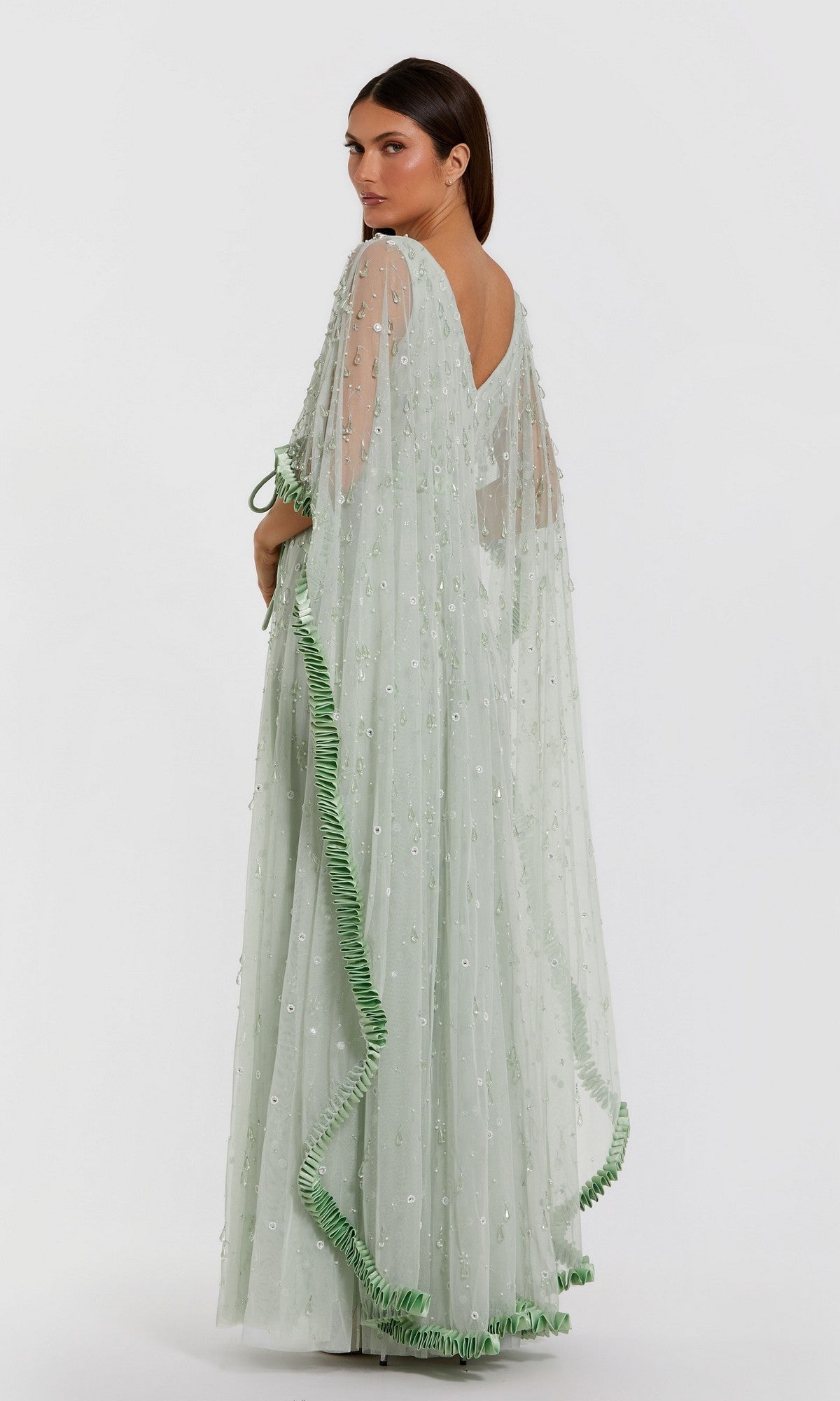 A woman stands against a white background, looking over her shoulder while wearing the Mac Duggal 94294 Long Formal Dress—a light green sheer gown featuring embroidered details and ruffled trim.