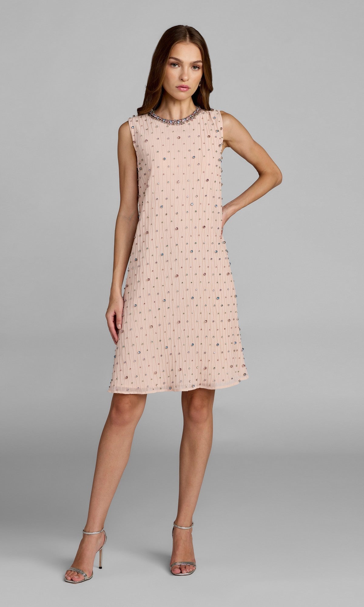 A woman stands in a sleeveless, blush multi Mac Duggal 94354 knee length shift party dress, featuring sparkling gem embellishments and paired with silver high-heeled sandals, against a plain gray background.