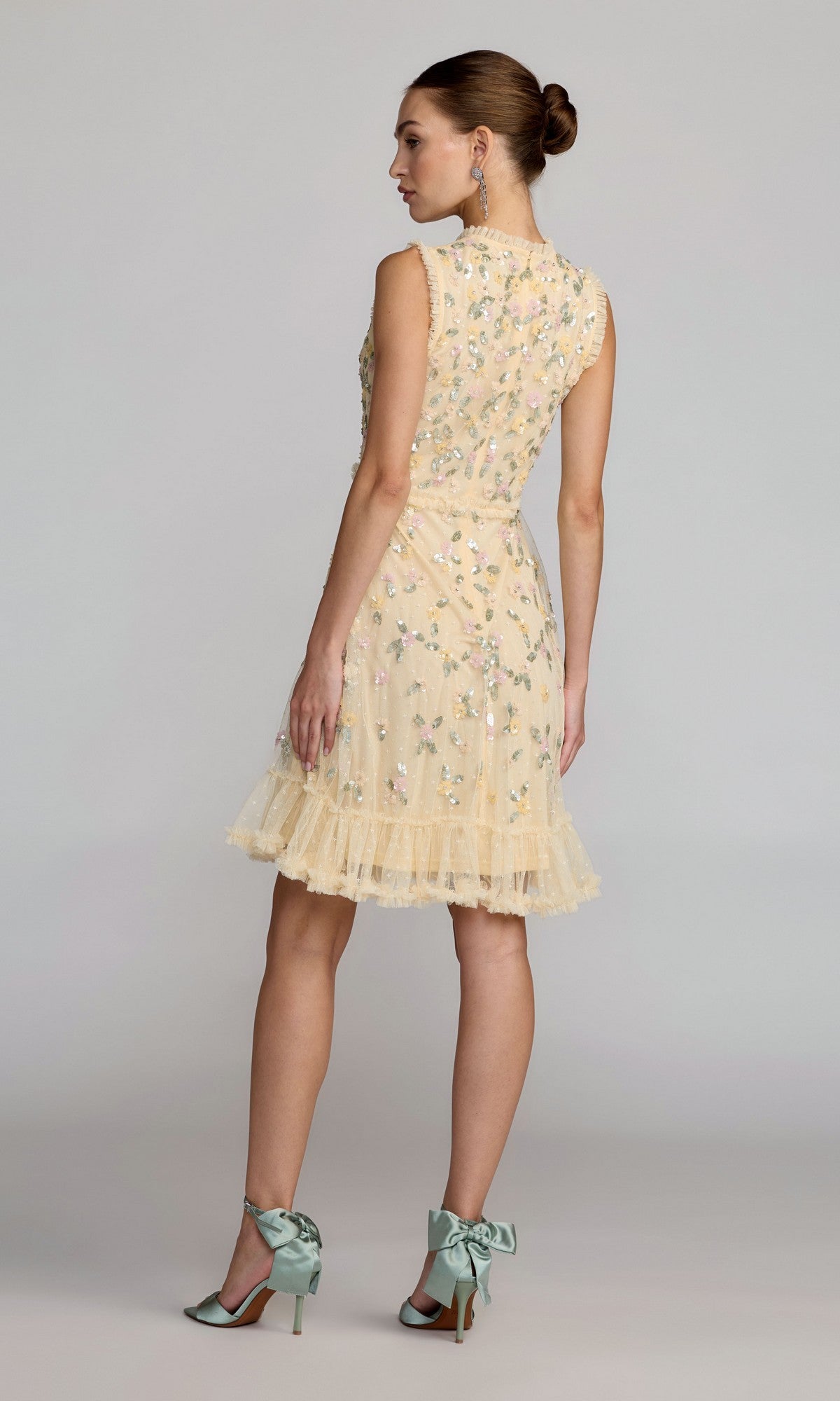 A woman stands in profile wearing the Mac Duggal 9474 Knee-Length Floral-Sequin Dress, featuring a lemon yellow hue, floral embroidery, ruffled hem, and paired with mint green heels tied with bows.