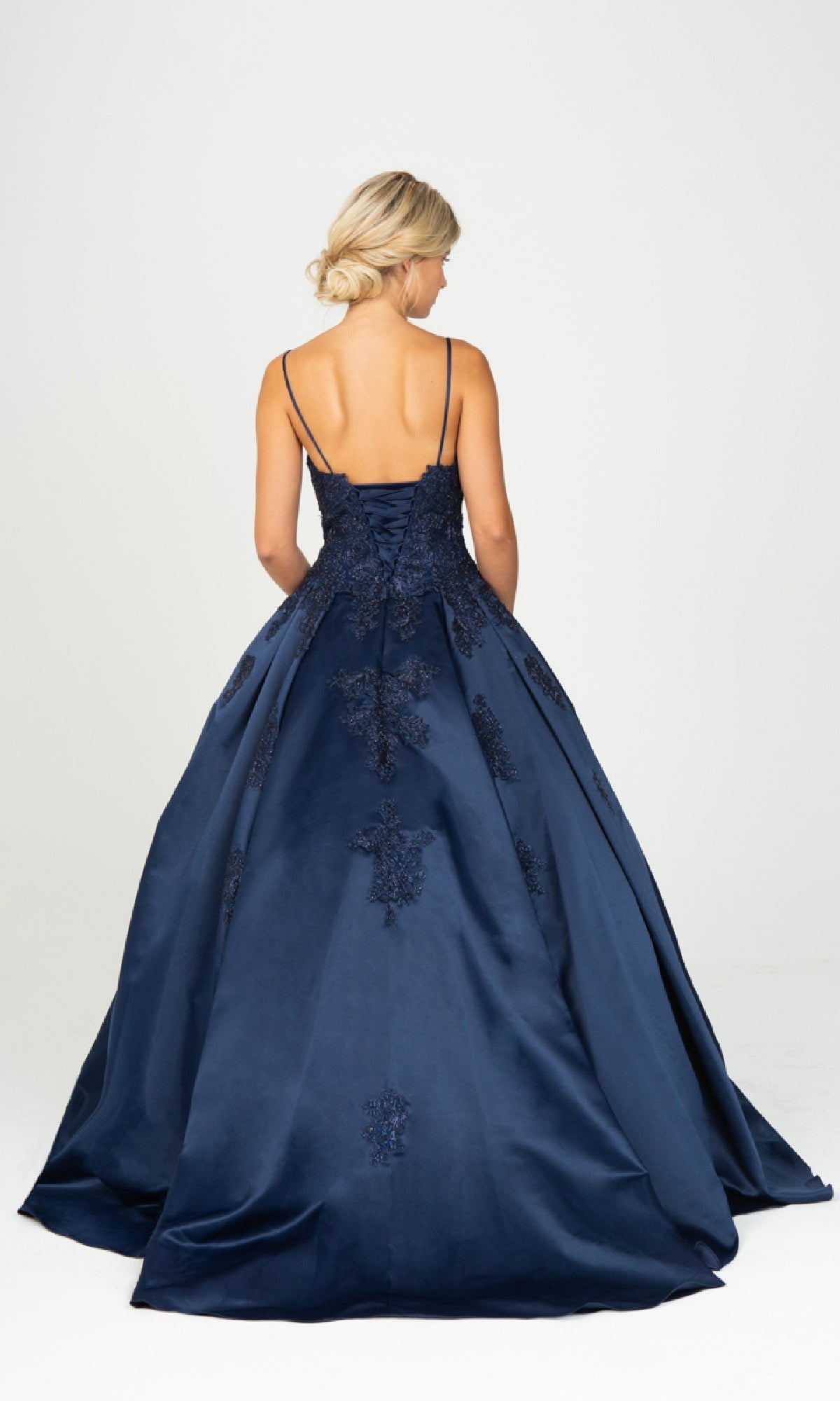 A woman stands facing away, wearing the Classic Long Prom Ball Gown: Eureka 9500—a navy blue floor-length dress with lace details, thin straps, and a lace-up back. Her blonde hair is styled in a low updo.