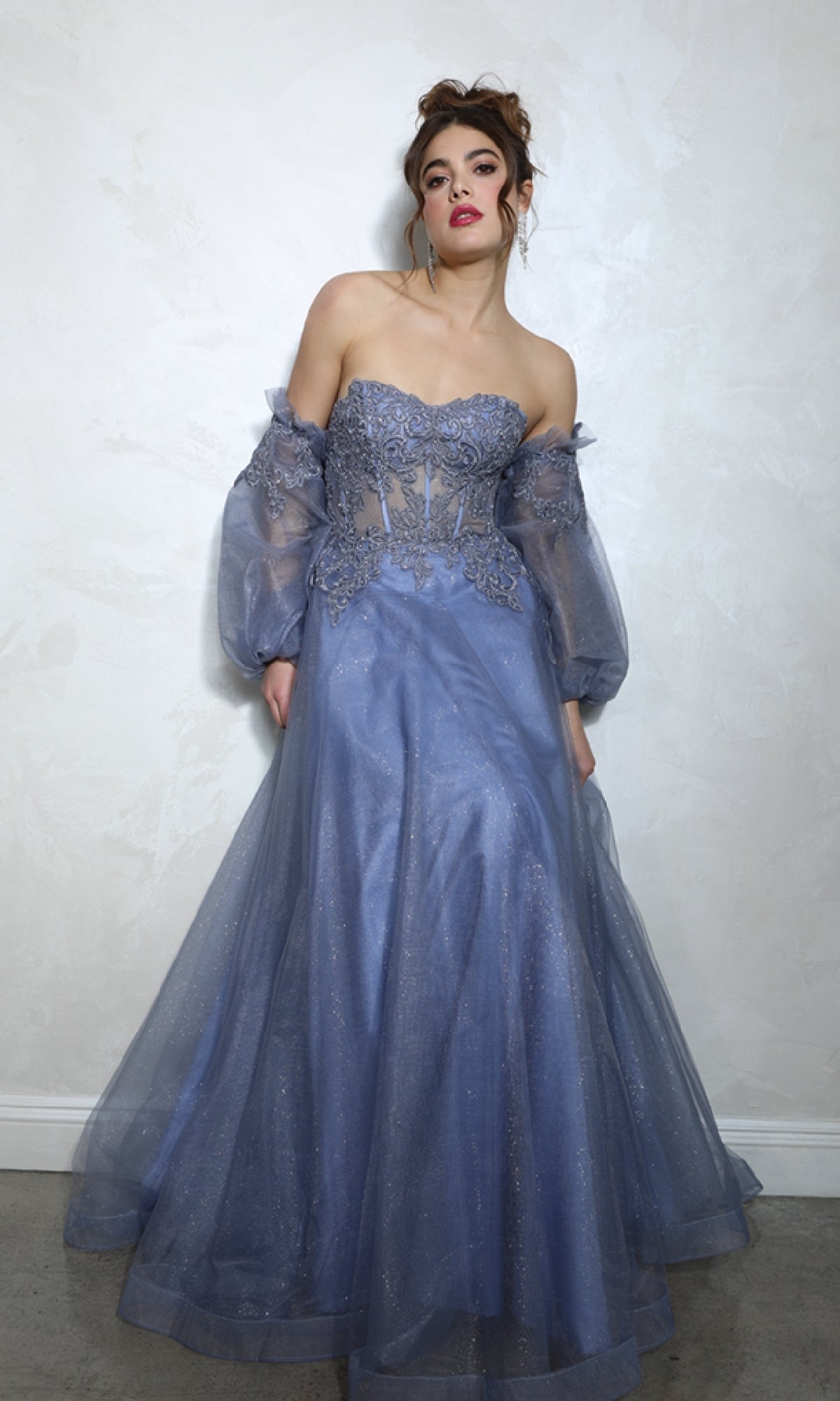 A woman stands by a light wall in the Eureka 9505G: a lavender, strapless prom ball gown with long sheer puffed sleeves and lace bodice details.