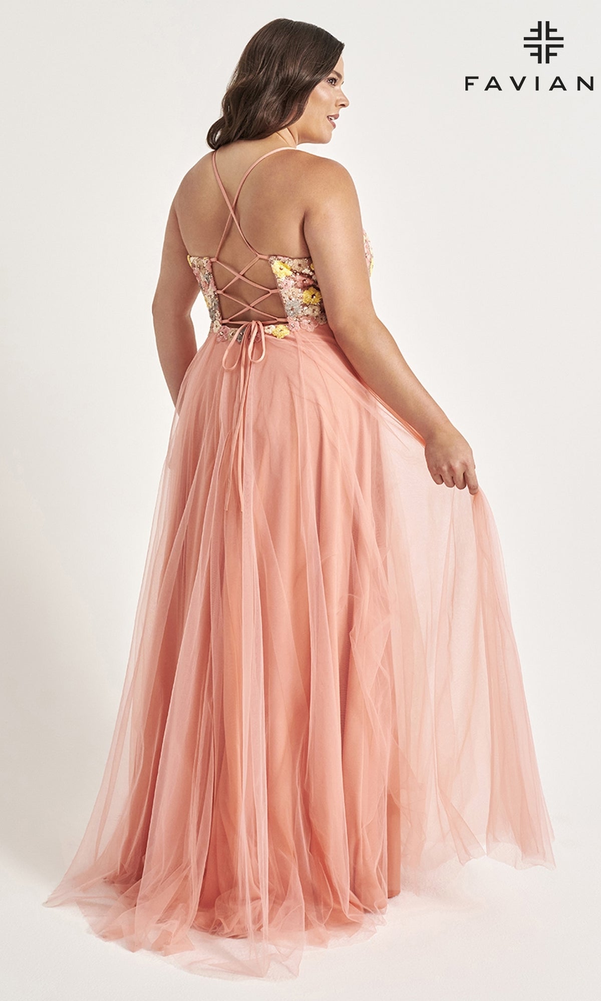 A woman wears the Faviana Floral Plus-Size Long Prom Dress 9557, featuring a peach hue, floor-length tulle skirt, lace-up back, thin straps, and floral detailing on the bodice. The