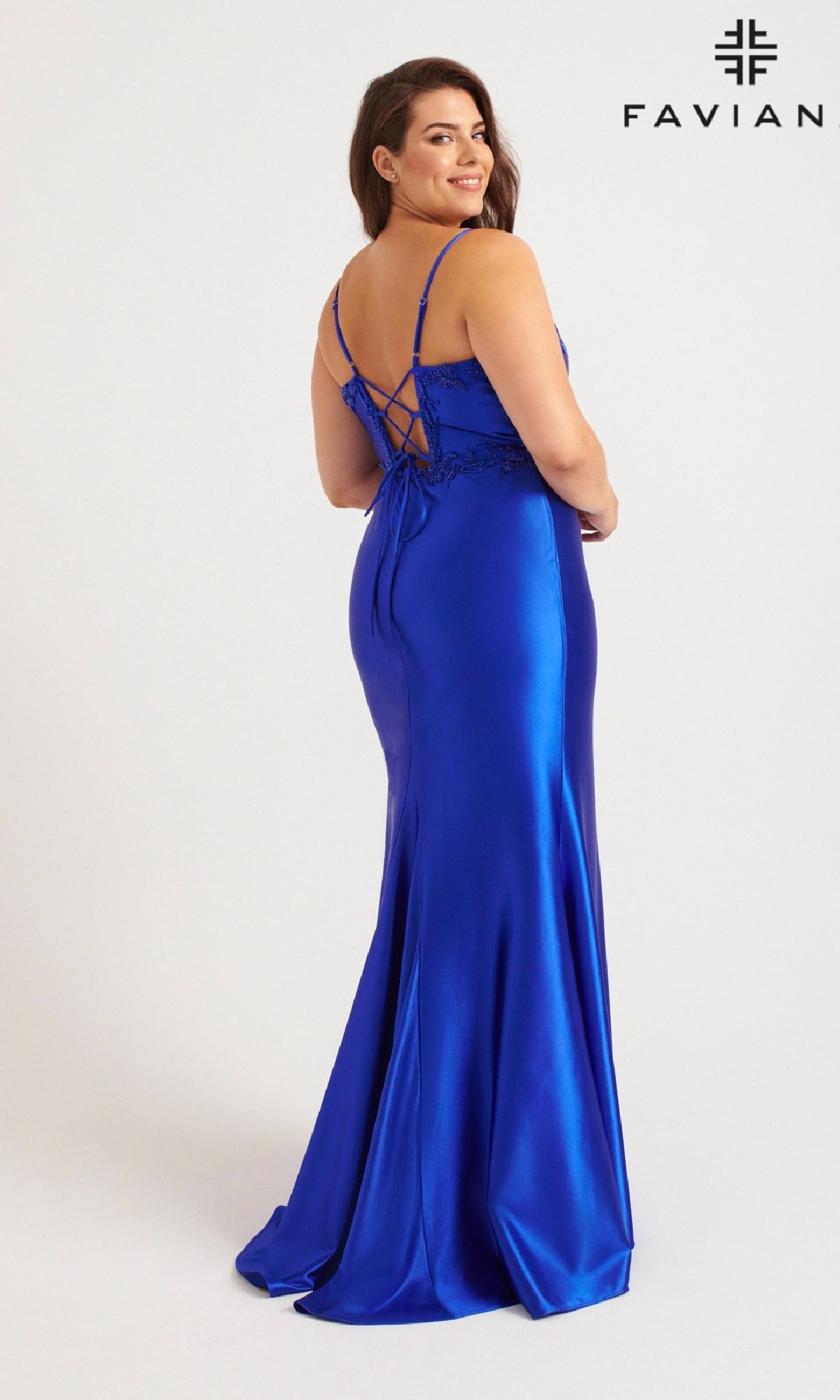 A woman in a bright blue satin long plus-size prom dress, Faviana 9562, with a beaded bodice and lace-up back stands facing away, looking over her shoulder. The Faviana logo appears in the upper right corner.