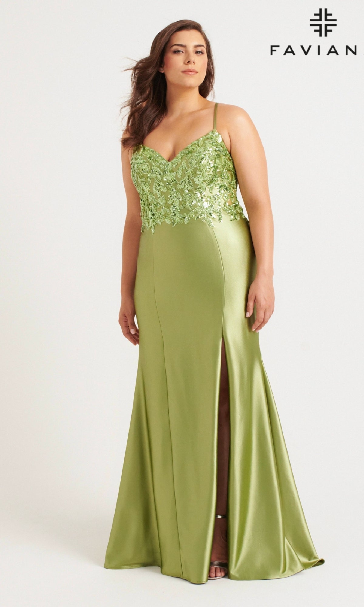 A woman stands against a plain background in the Faviana 9563 Long Plus-Size Prom Dress, a light green satin gown with floral lace details, thin straps, and a thigh-high slit. The Faviana logo appears at the top right corner.