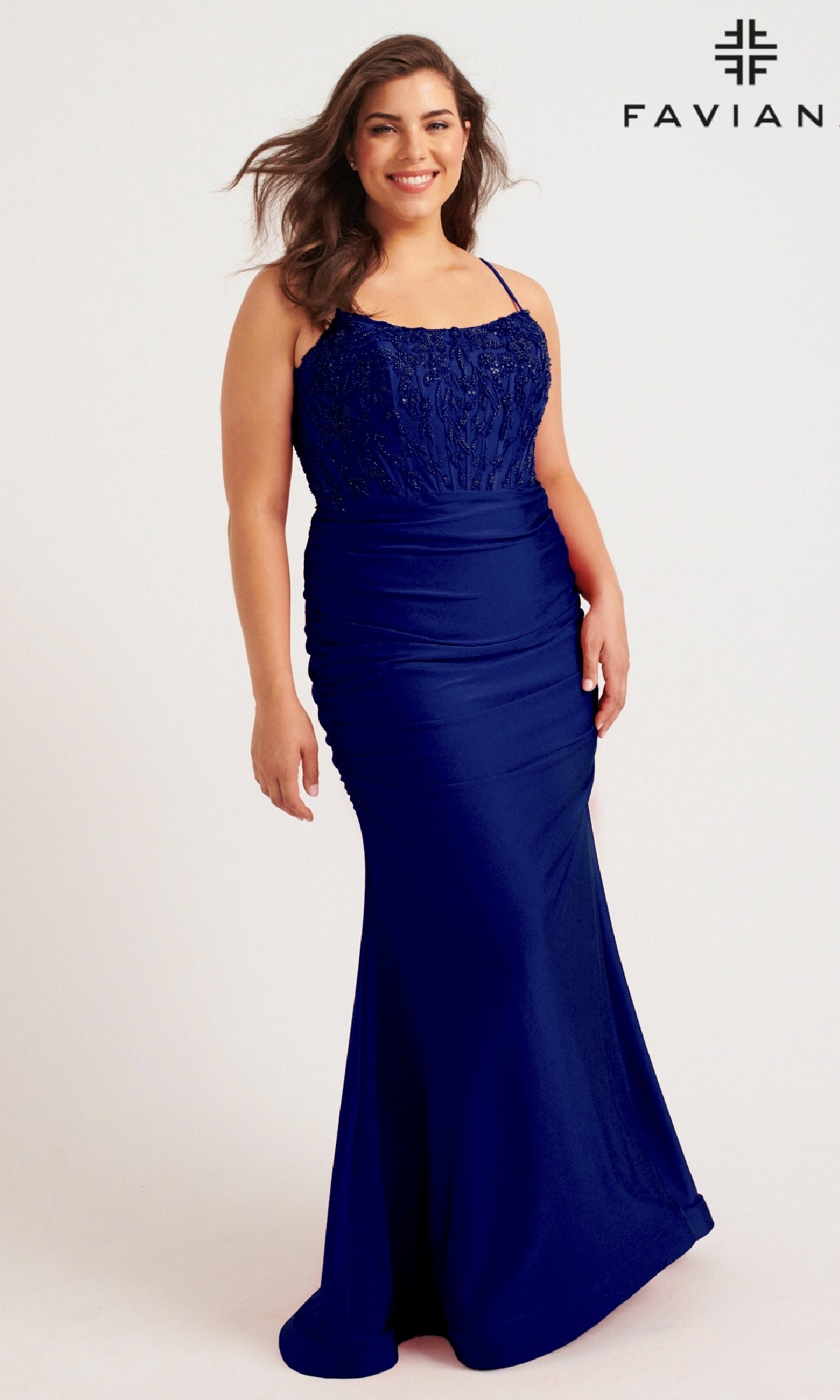 A woman models the Faviana 9569, a long plus-size prom dress in royal blue with lace and beaded corset bodice, standing against a plain background. The