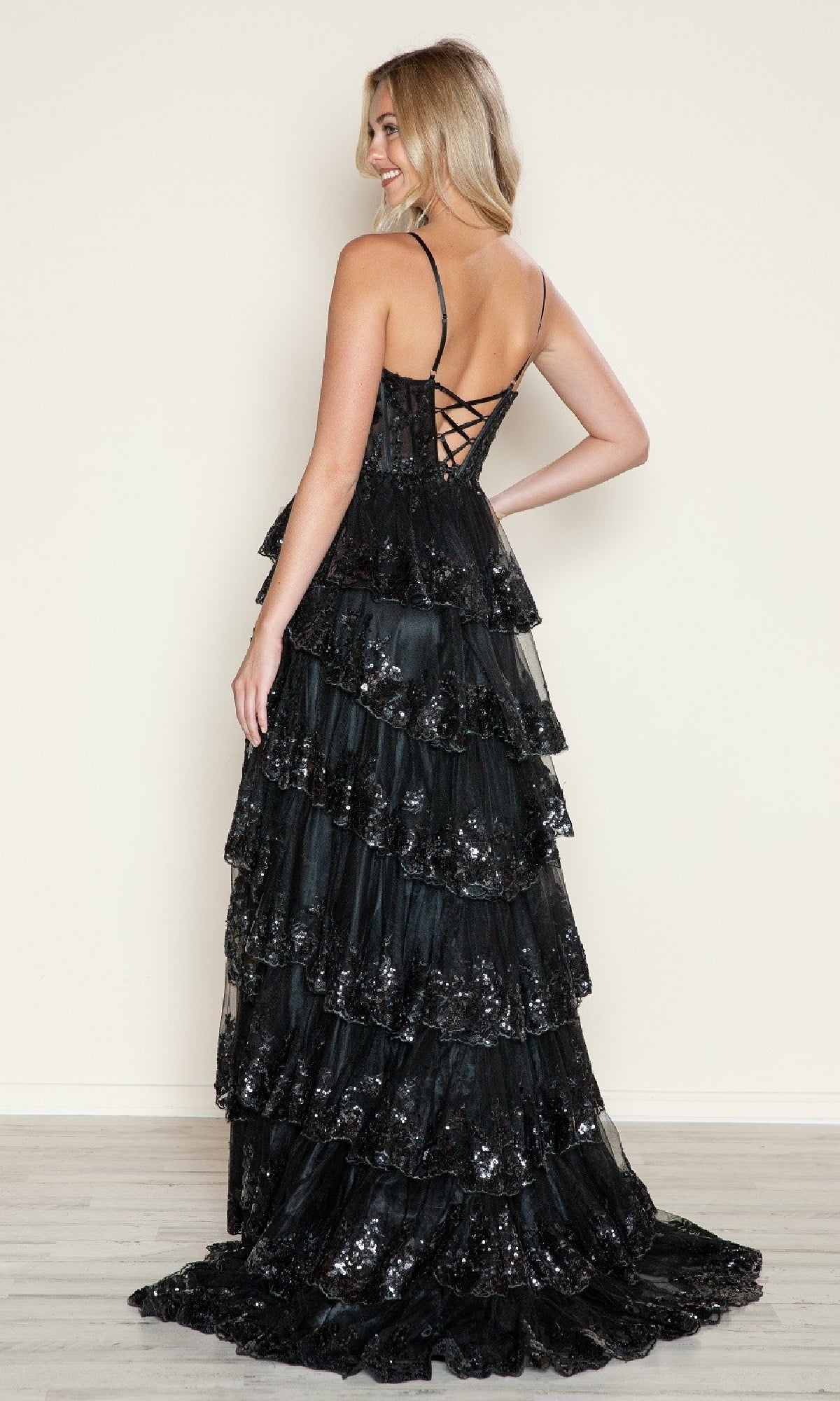 A woman stands on a light floor facing away, wearing the Poly USA 9606 Long Prom Dress—black, tiered A-line with sequins, thin straps, and a lace-up back.