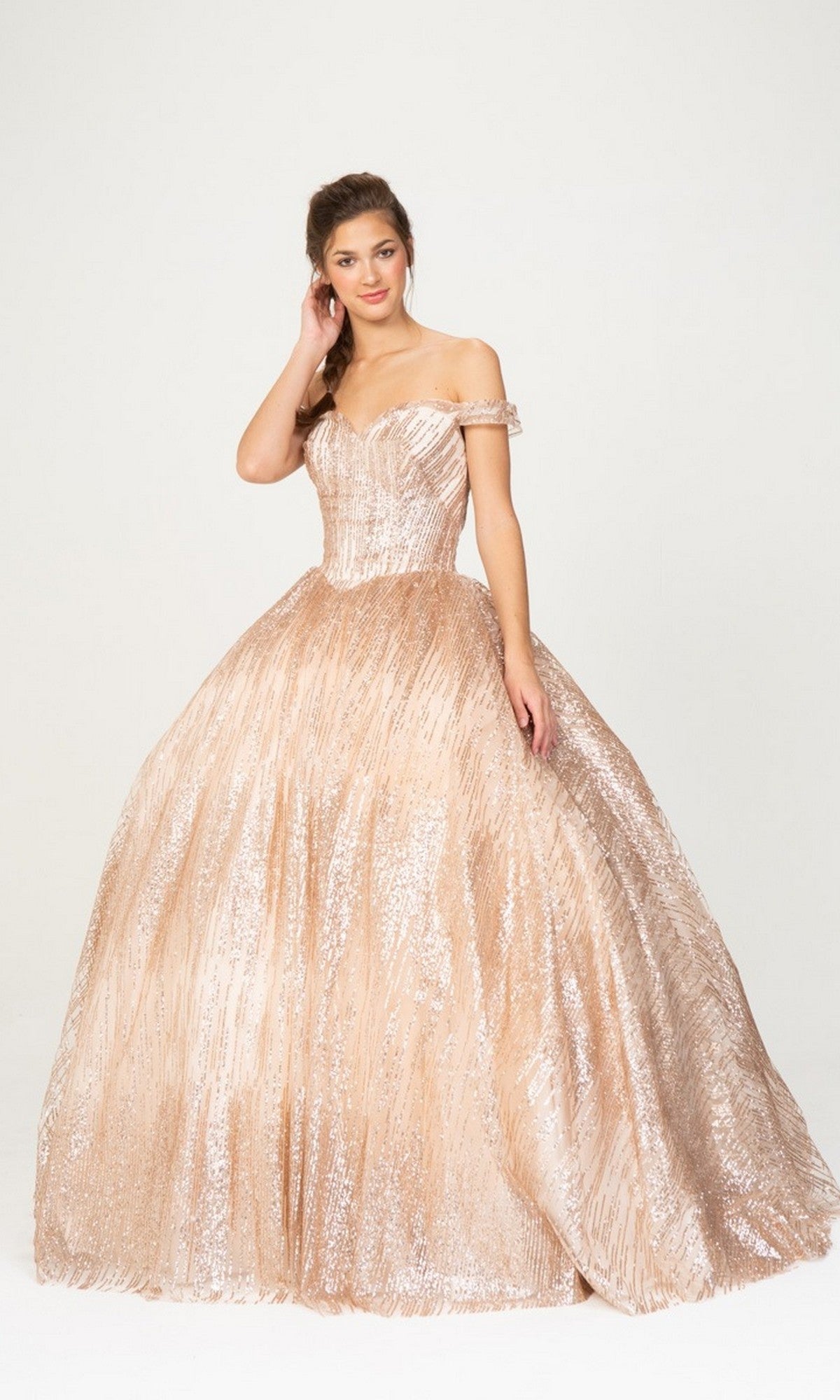 A woman is wearing the Quinceanera Dress 9616 by Eureka, an off-the-shoulder, champagne-colored ball gown with a full, shimmery skirt, posing gracefully against a plain white background.