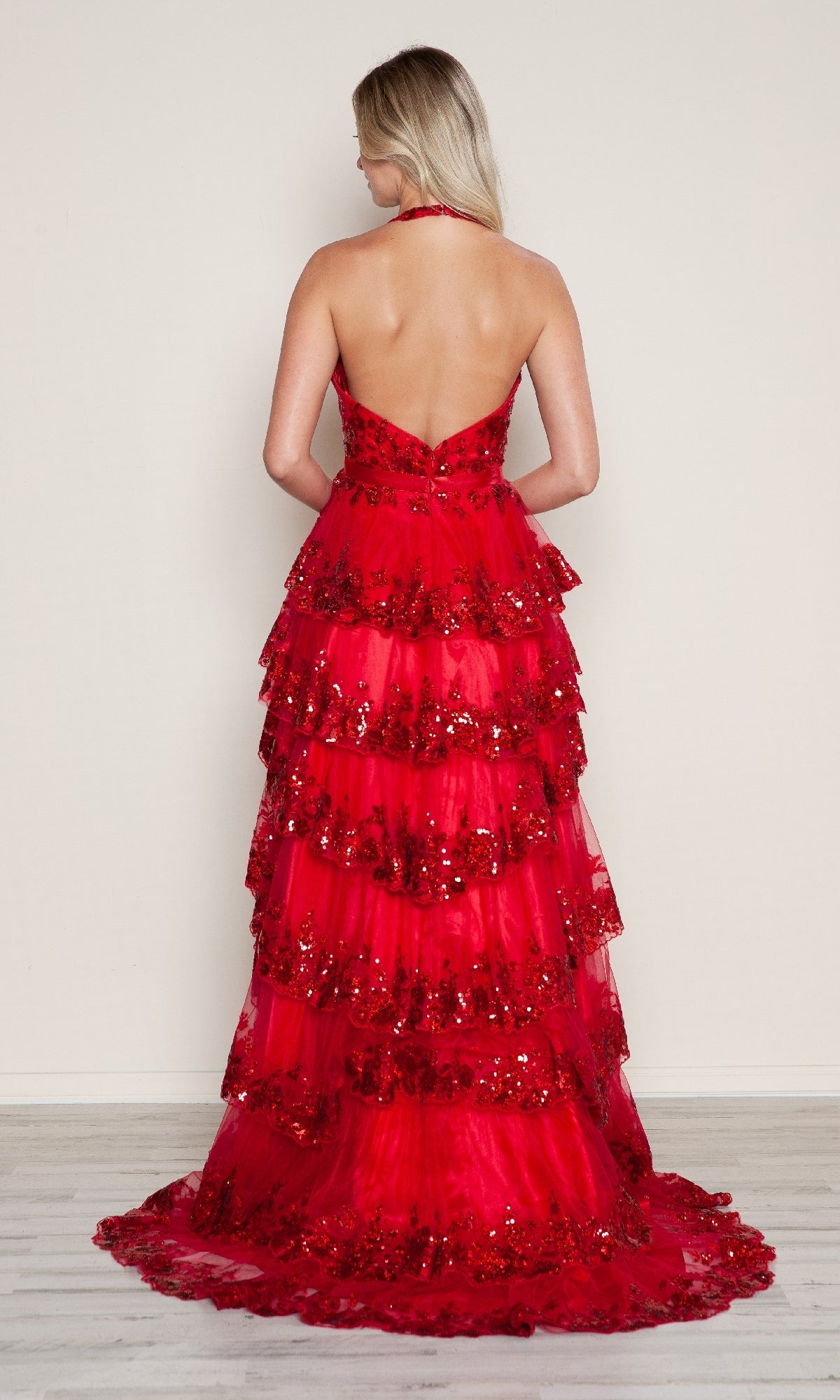 A blonde woman models the Poly USA 9618 long prom dress, featuring a halter neck, floor-length ruffled layers, and sequin embellishments, standing against a plain background.
