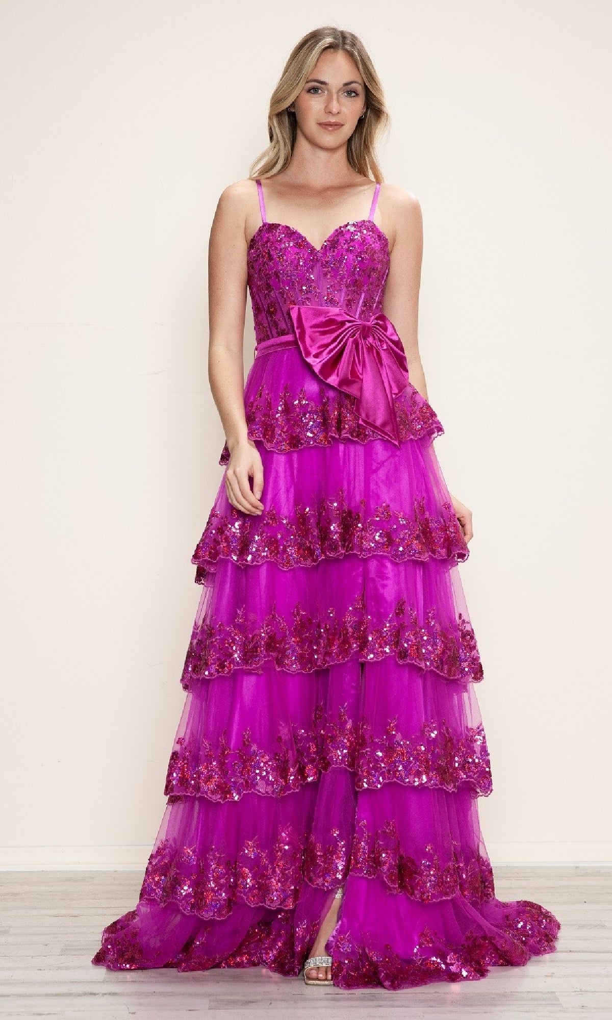 A woman stands against a plain background wearing the Poly USA 9626 long prom dress—a bright pink A-line gown with a tiered skirt, sequin embellishments, thin straps, sweetheart neckline, and a large waist bow.