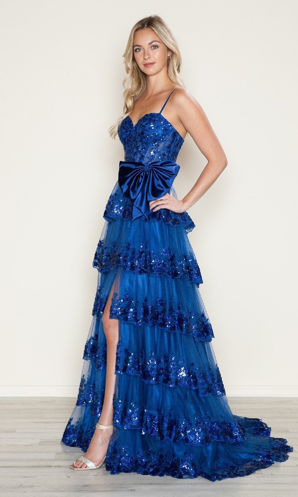 A woman stands against a plain wall wearing the Poly USA 9626 long prom dress, featuring an A-line silhouette, blue tiered skirt, sequin embellishments, thin straps, a large waist bow, and silver high heel sandals.