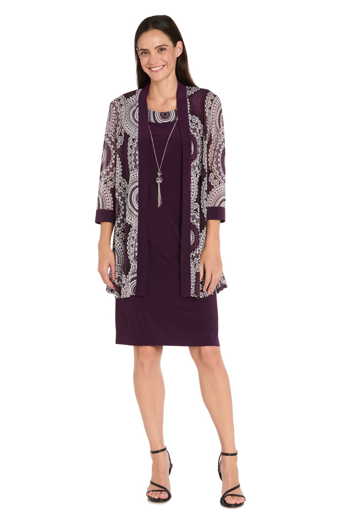 A woman wearing the Petite Two-Piece Short Party Dress: R&M Richards 9626P, featuring a short purple cocktail dress with a patterned sheer petite jacket, black sandals, and a long pendant necklace, smiles against a white background.