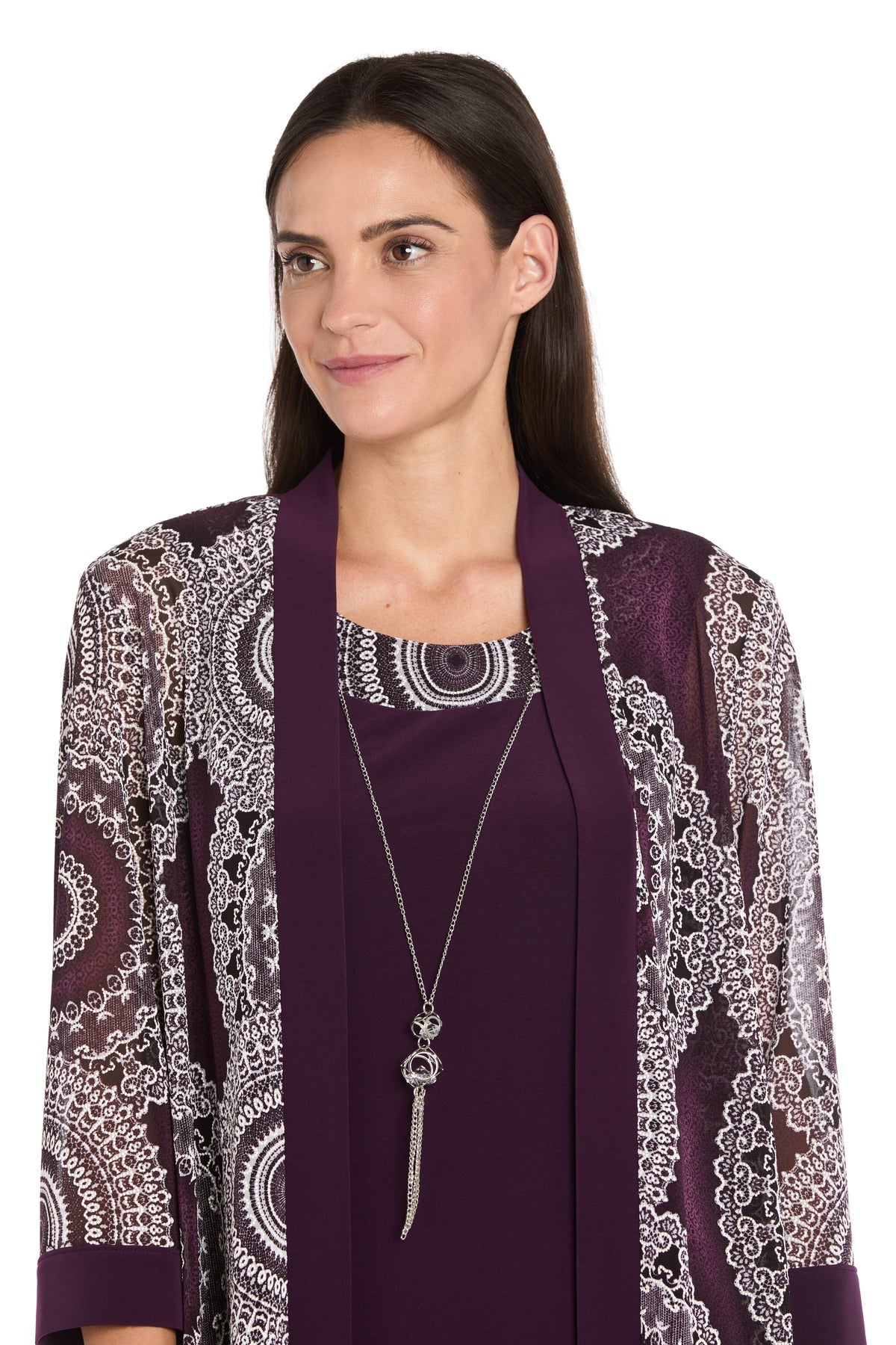 A woman with straight dark hair, facing slightly to the side and smiling, wears the R&M Richards 9626P Petite Two-Piece Short Party Dress in eggplant, paired with a long purple scarf and a silver pendant necklace.
