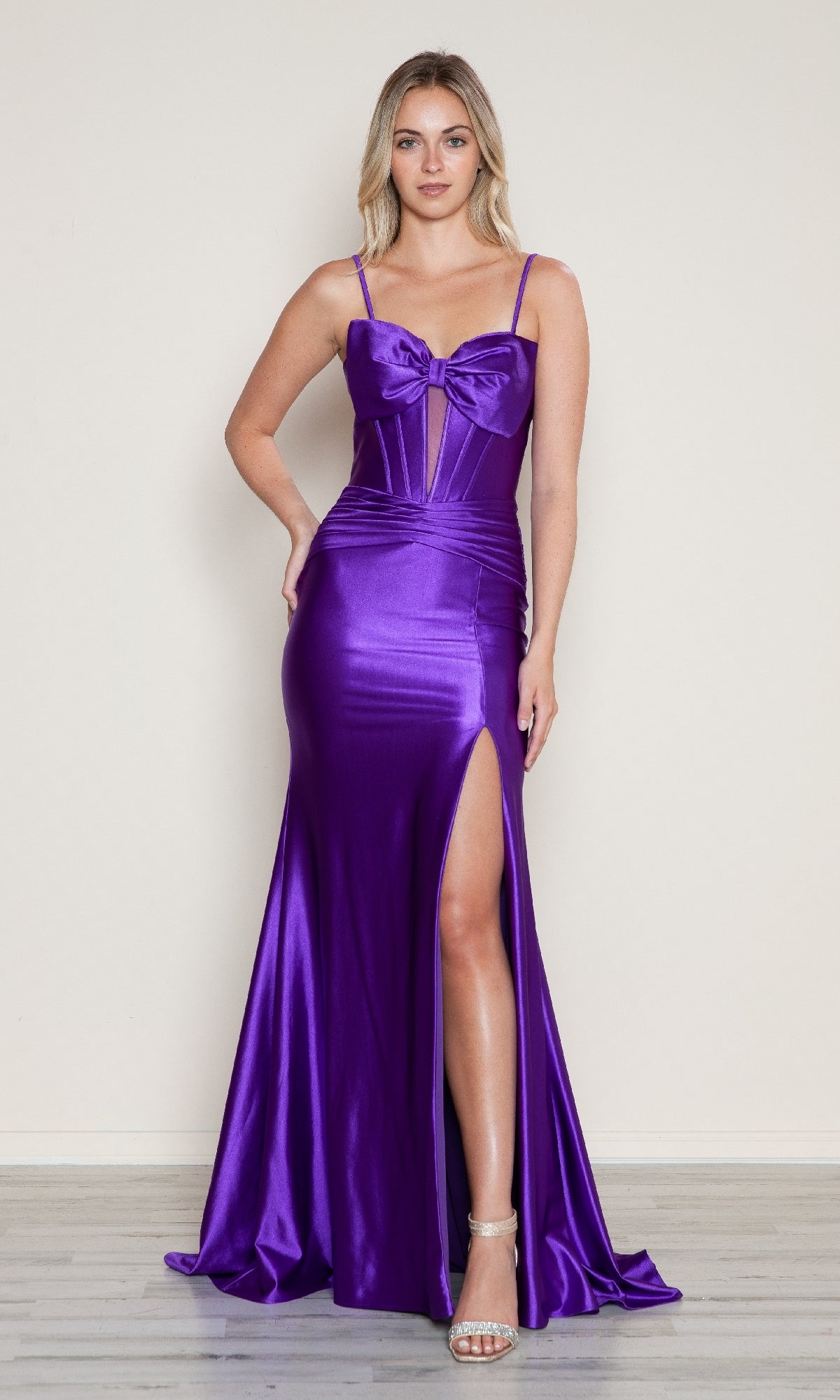 Woman wearing the Front-Bow Long Cut-Out Prom Dress (Poly USA 9644), a shiny purple evening gown with spaghetti straps, front bow detail, and high slit, paired with silver heels against a plain background.
