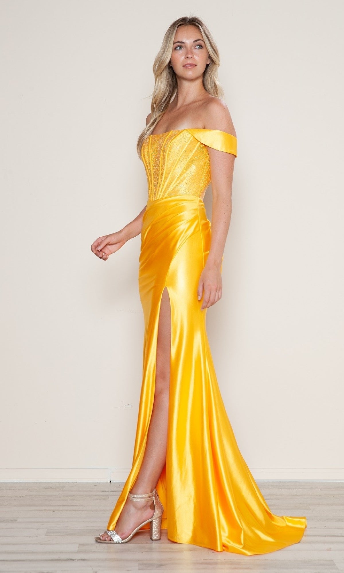 A woman stands indoors wearing the Off-Shoulder Long Corset Prom Dress: Poly USA 9656 in bright yellow, featuring a beaded corset bodice, thigh-high slit, and paired with silver high-heeled sandals.