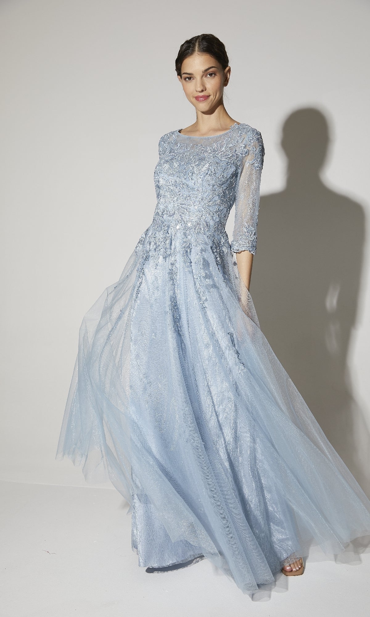 A woman models the Eureka 9660, a flowy long sleeve formal dress with a light blue lace bodice and sheer sleeves, posed against a plain background.
