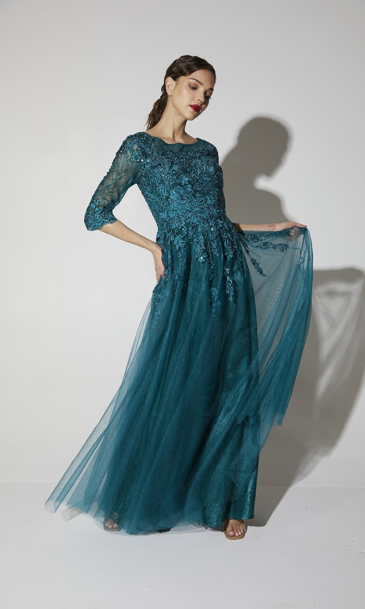 Woman wearing the Flowy Long Sleeve Long Formal Dress: Eureka 9660, showcasing a teal lace bodice, three-quarter sleeves, and a flowing tulle skirt against a plain white background.