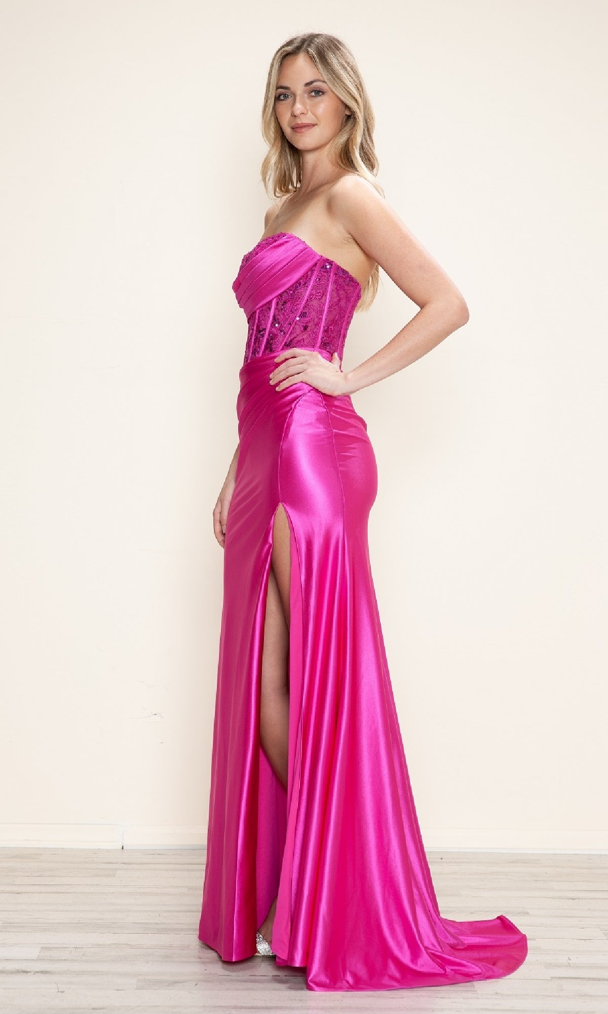 A woman poses sideways wearing the Strapless Long Corset Prom Gown: Poly USA 9688—a bright pink, strapless long dress with a high slit and sheer lace corset bodice—on a light wood floor against an off-white wall.