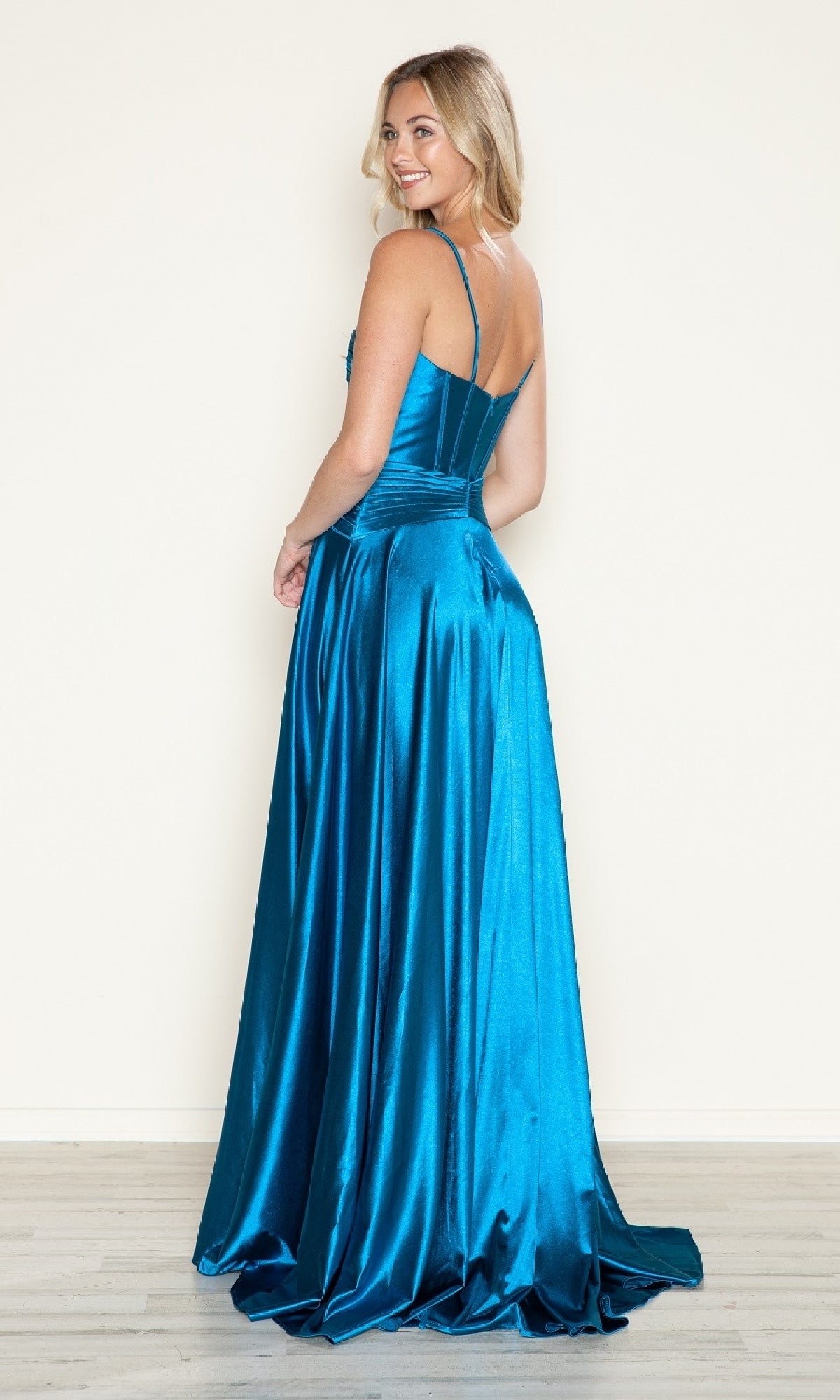 A woman in the Cut-Out Long A-Line Prom Dress: Poly USA 9700, a sleeveless blue satin gown, stands on a light wood floor, facing away and smiling over her shoulder.
