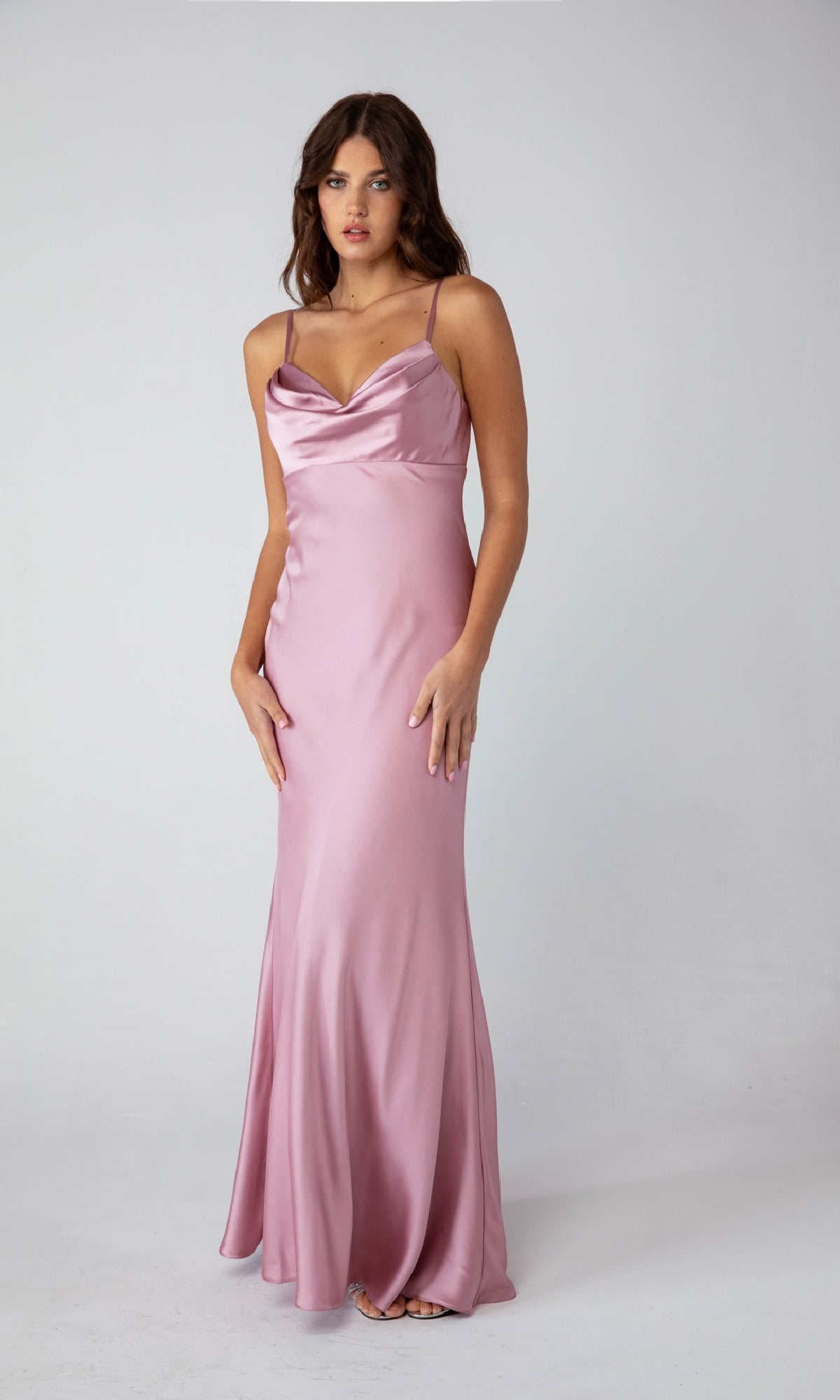 A woman poses in a studio wearing the Eureka 9711 Cowl-Neck Long Satin Prom Dress, a sleeveless, floor-length evening gown in light pink with a draped neckline.