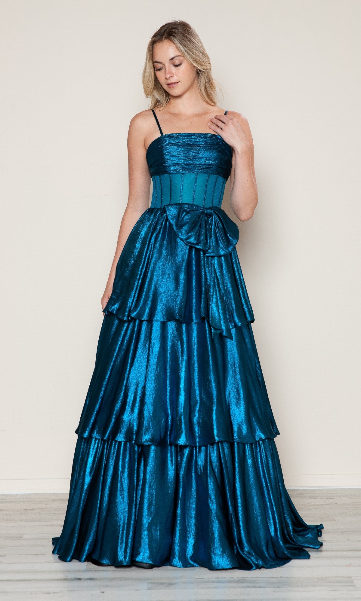 A woman stands indoors wearing the Long Prom Dress: Poly USA 9718, featuring a sleeveless teal design with thin straps, a fitted bodice, bow at the waist, and tiered A-line skirt.