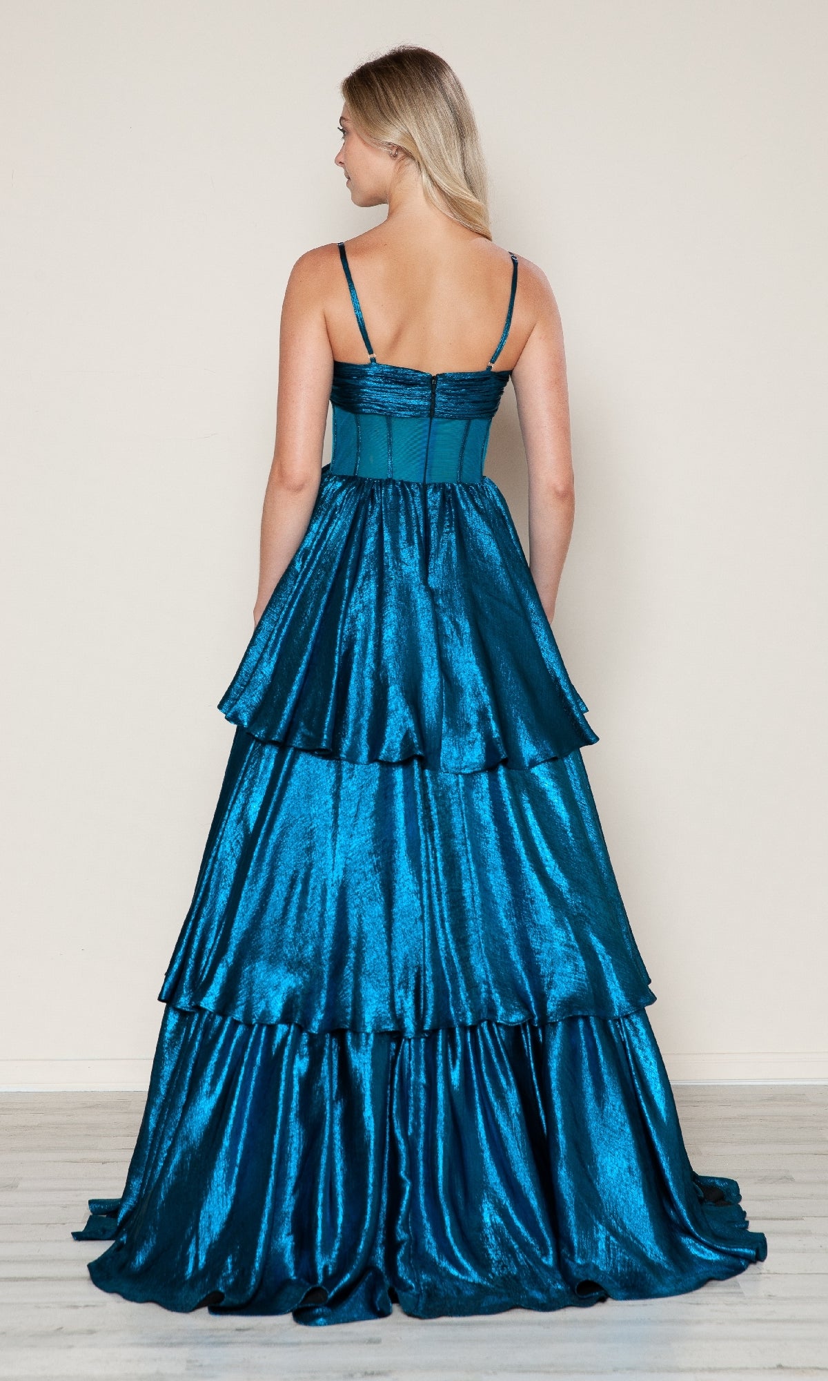 A woman with blonde hair is seen from the back wearing the Poly USA 9718 long prom dress, featuring shiny teal metallic fabric, thin straps, and a tiered A-line skirt, standing against a plain light-colored wall.