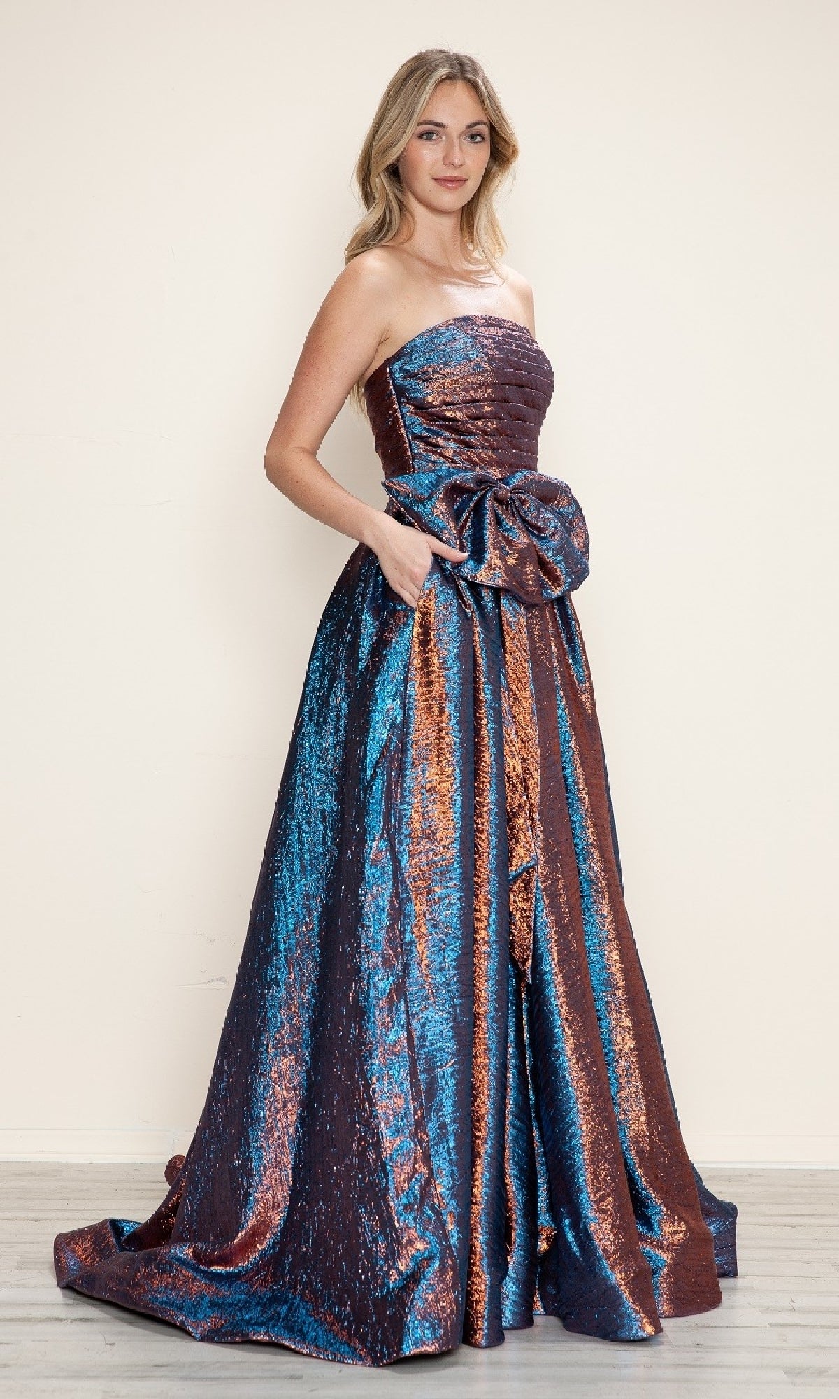 A woman wears the Side-Bow Strapless Prom Gown (Poly USA 9720), a floor-length evening dress with blue and copper metallic stripes, a strapless neckline, and a large bow at the waist.