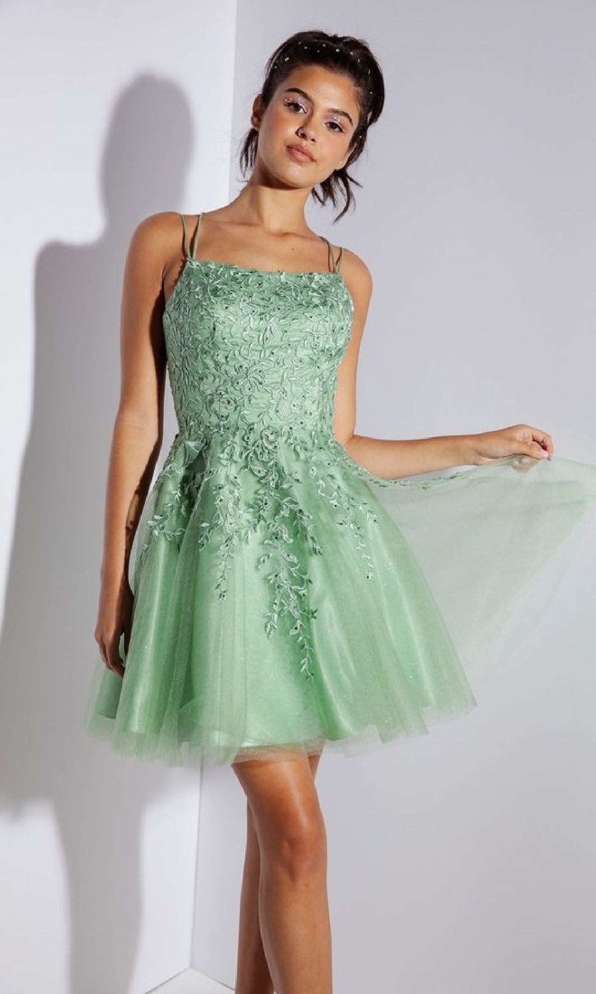 A woman stands indoors wearing the Eureka 9727 Lace-Up Short Prom Dress, a light green, sleeveless A-line party dress with floral embroidery and a tulle skirt.