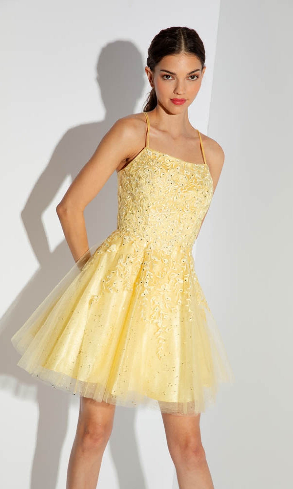 A woman with dark braided hair stands against a light background, wearing the Eureka 9727 Lace-Up Short Prom Dress—a yellow, sleeveless, floral lace cocktail dress with embroidery and tulle—looking at the camera.