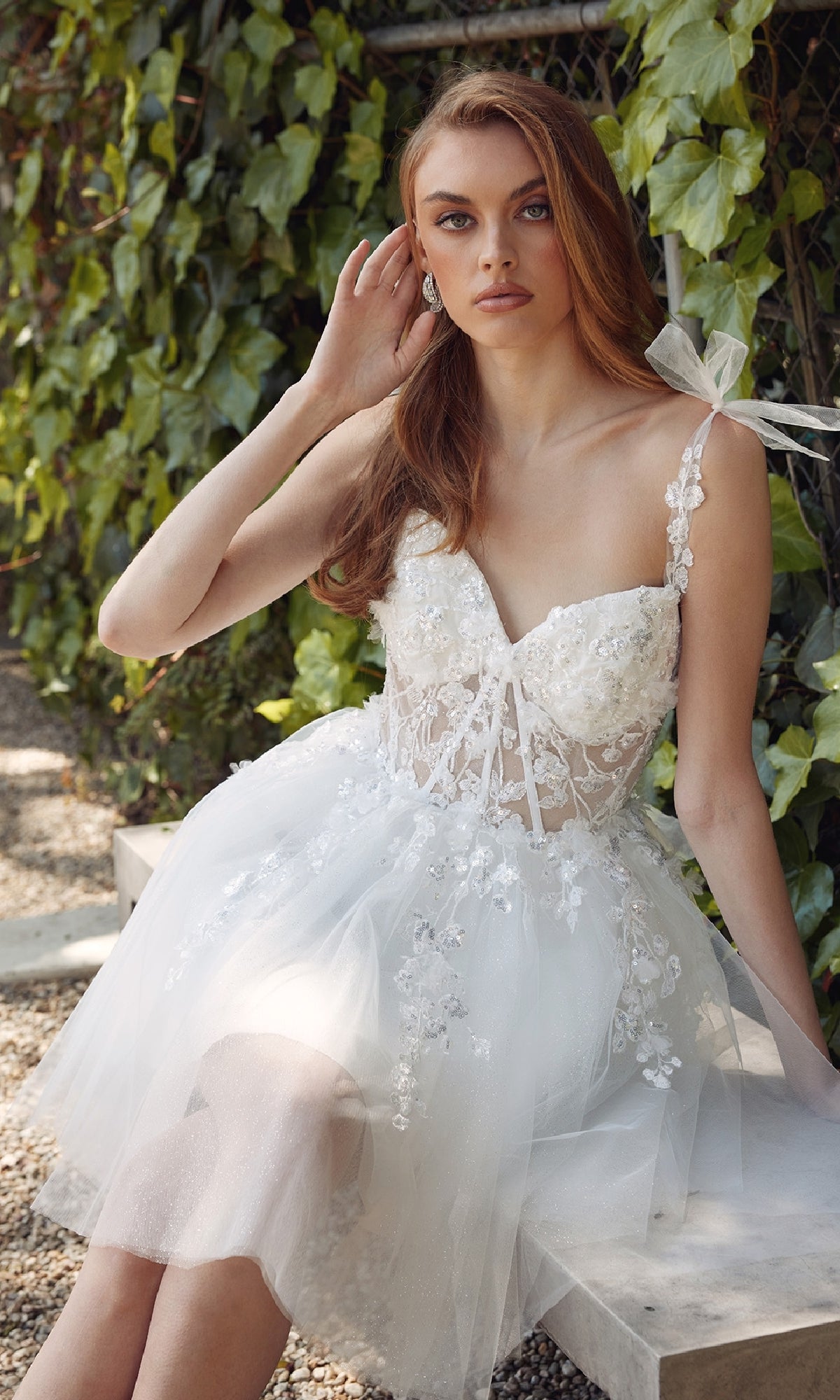 A woman wearing the Juliet JT972LW short white bow hoco dress sits on a stone bench outdoors, framed by lush green ivy leaves.