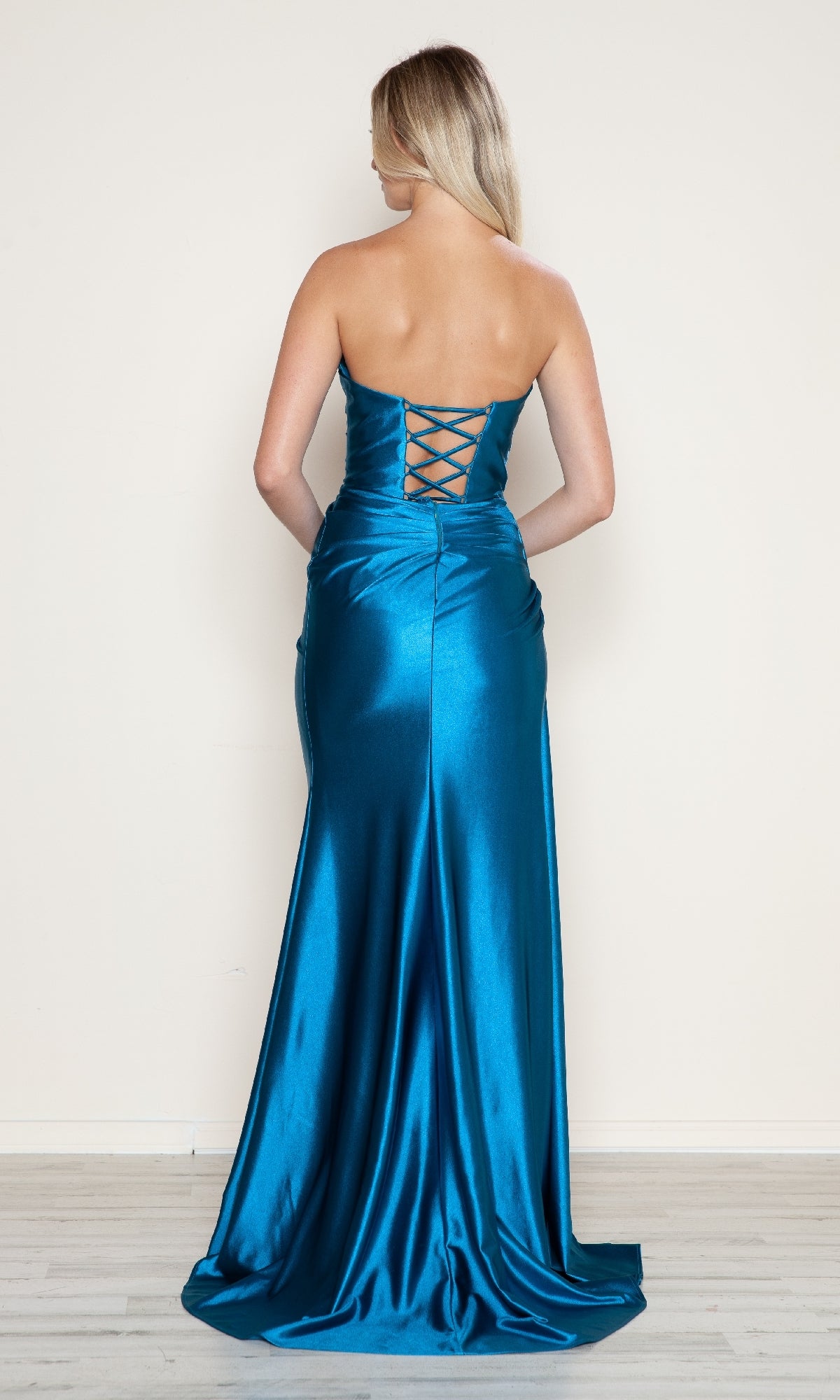 A woman faces away from the camera in the Strapless Side-Sash Long Prom Gown: Poly USA 9734, highlighting its lace-up back and teal satin fabric against a simple light background.