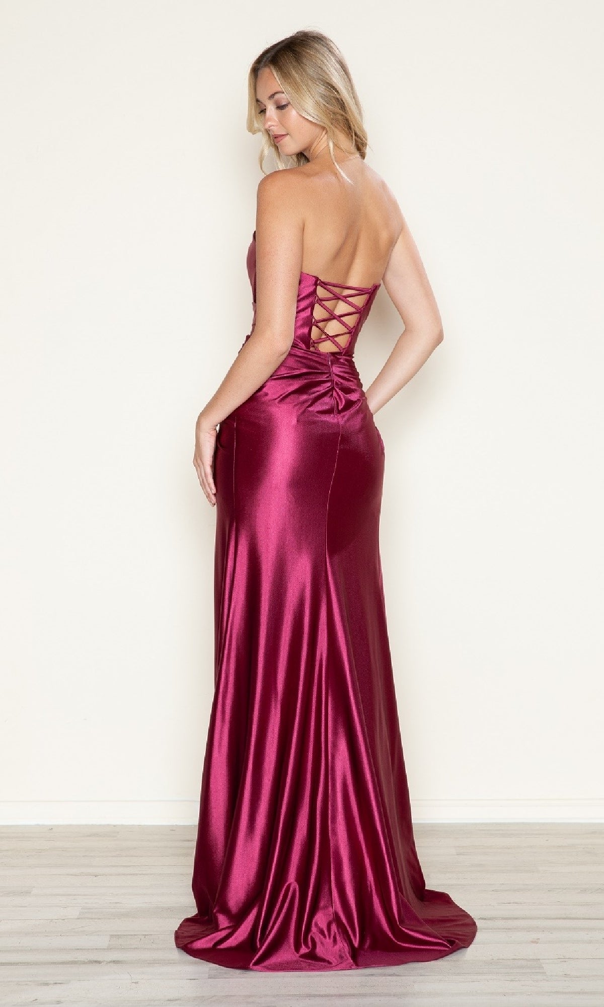 A woman faces away on a light wood floor wearing the Strapless Side-Sash Long Prom Gown: Poly USA 9734, a shiny burgundy satin evening dress with a sweetheart neckline and lace-up back—an elegant choice for under $200.