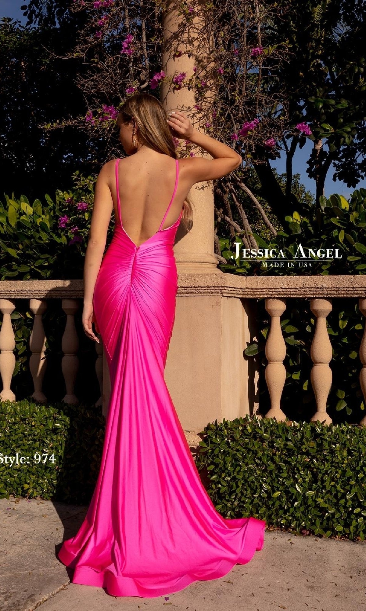 A woman stands outdoors with her back to the camera, wearing the bright pink Jessica Angel 974 Open-Back Long Prom Dress with a mermaid skirt near a stone railing and blooming flowers.