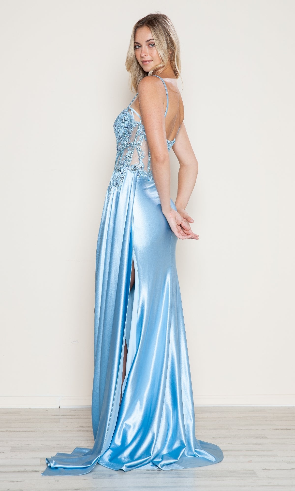 A woman models the Poly USA 9740 long prom dress, featuring light blue satin, an embroidered sheer corset bodice, and a high leg slit, while standing against a plain light-colored wall.