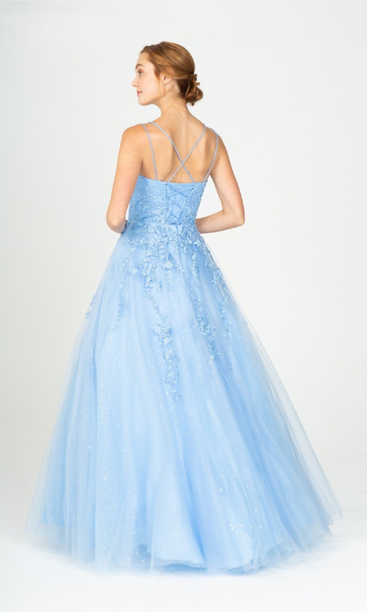 A woman stands facing away, wearing the Eureka CC0240 Lace-Up Long Lace Prom Ball Gown, featuring a fitted strappy bodice and a flowing embroidered tulle skirt in light blue.