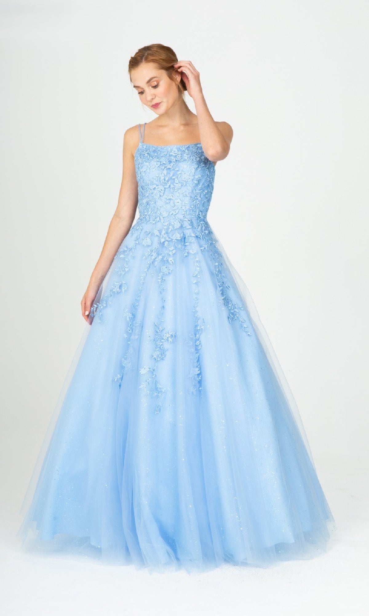A woman stands in the Eureka CC0240 Lace-Up Long Lace Prom Ball Gown, a light blue A-line dress with floral embellishments, thin straps, and a delicate lace-up back, set against a plain white background.