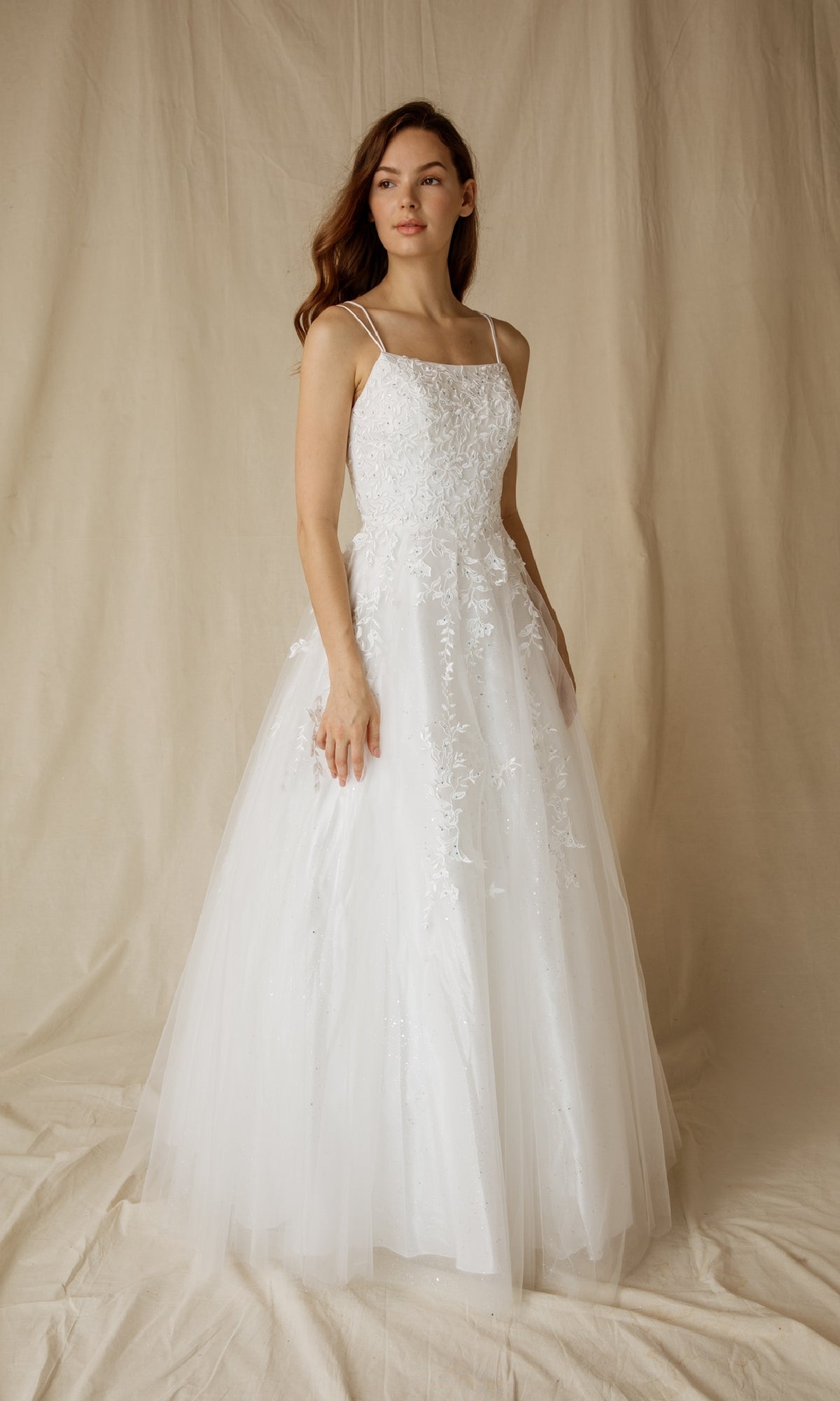 A woman in the Eureka CC0240 Lace-Up Long Lace Prom Ball Gown stands against a beige fabric backdrop, looking slightly to the side. The gown features white lace, sleeveless design, lace detailing, and a lace-up back.