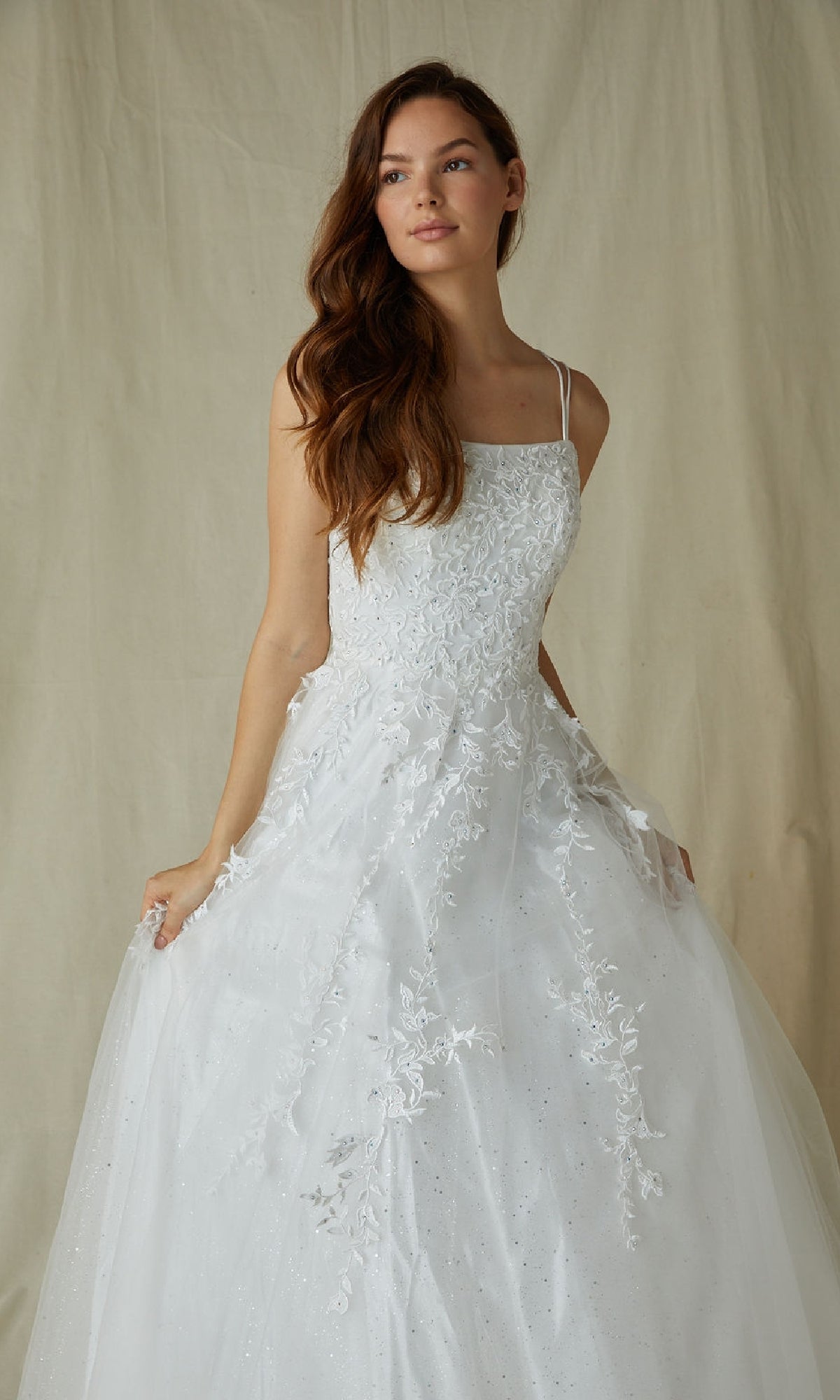 A woman in the Eureka CC0240 Lace-Up Long Lace Prom Ball Gown, featuring white floral embroidery, a tulle skirt, and lace-up back, poses against a beige fabric backdrop.