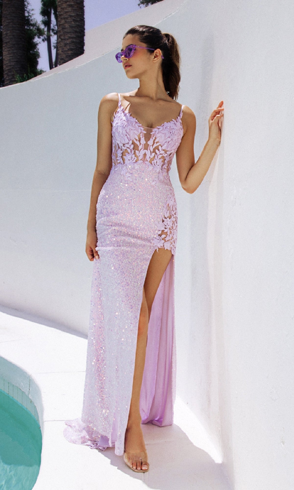 A woman stands by a white wall near a pool, wearing sunglasses and the Eureka 9790 long prom dress—a sleeveless lilac purple sequin gown with lace details, corset bodice, and high slit.