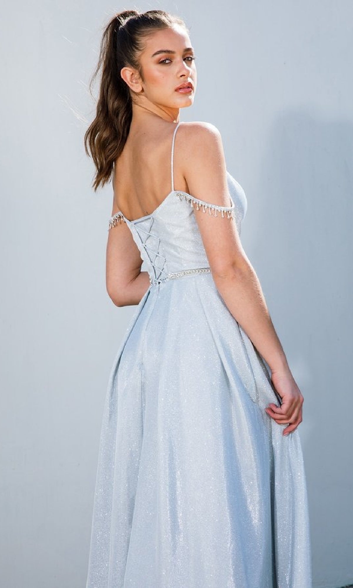 A young woman with long brown hair in a ponytail wears the Eureka 9800 Lace-Up Long Shimmer Prom Dress, a light blue, off-the-shoulder A-line gown with jeweled chains and a lace-up back, standing against a plain background.