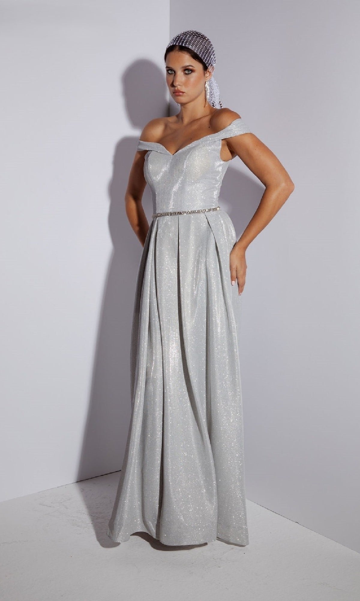 A woman models the Eureka 9808 Off-Shoulder Long Glitter Prom Dress—a silver gown with jeweled belt and netted headpiece—posing elegantly against a plain white background.