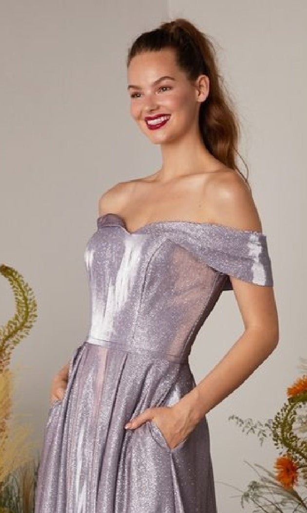 A woman smiles in front of a flower-adorned backdrop while wearing the Eureka 9809 Off-Shoulder Long Glitter Prom Dress, a shimmery lavender evening gown with pockets.