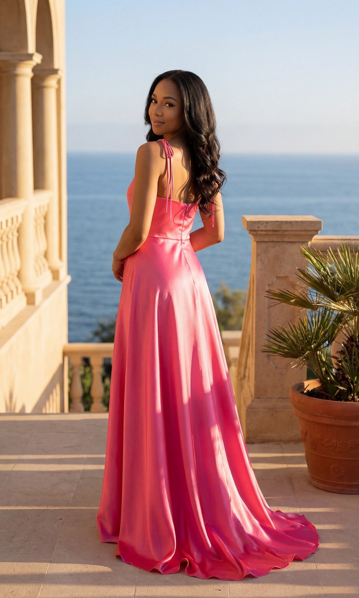 A woman in the Chicas 9811 long prom dress, featuring pink satin and an elegant leg slit, stands on a terrace with stone railings overlooking the sea on a sunny day.
