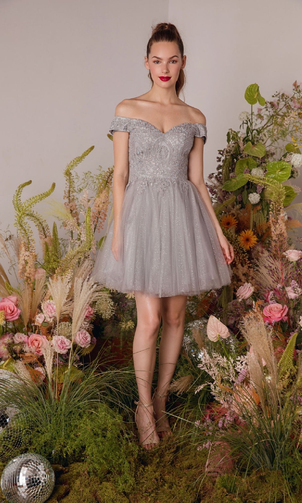 A woman stands on grass, surrounded by flowers, greenery, and two disco balls, wearing the Eureka 9833 Off-the-Shoulder Short Hoco Dress in light gray.