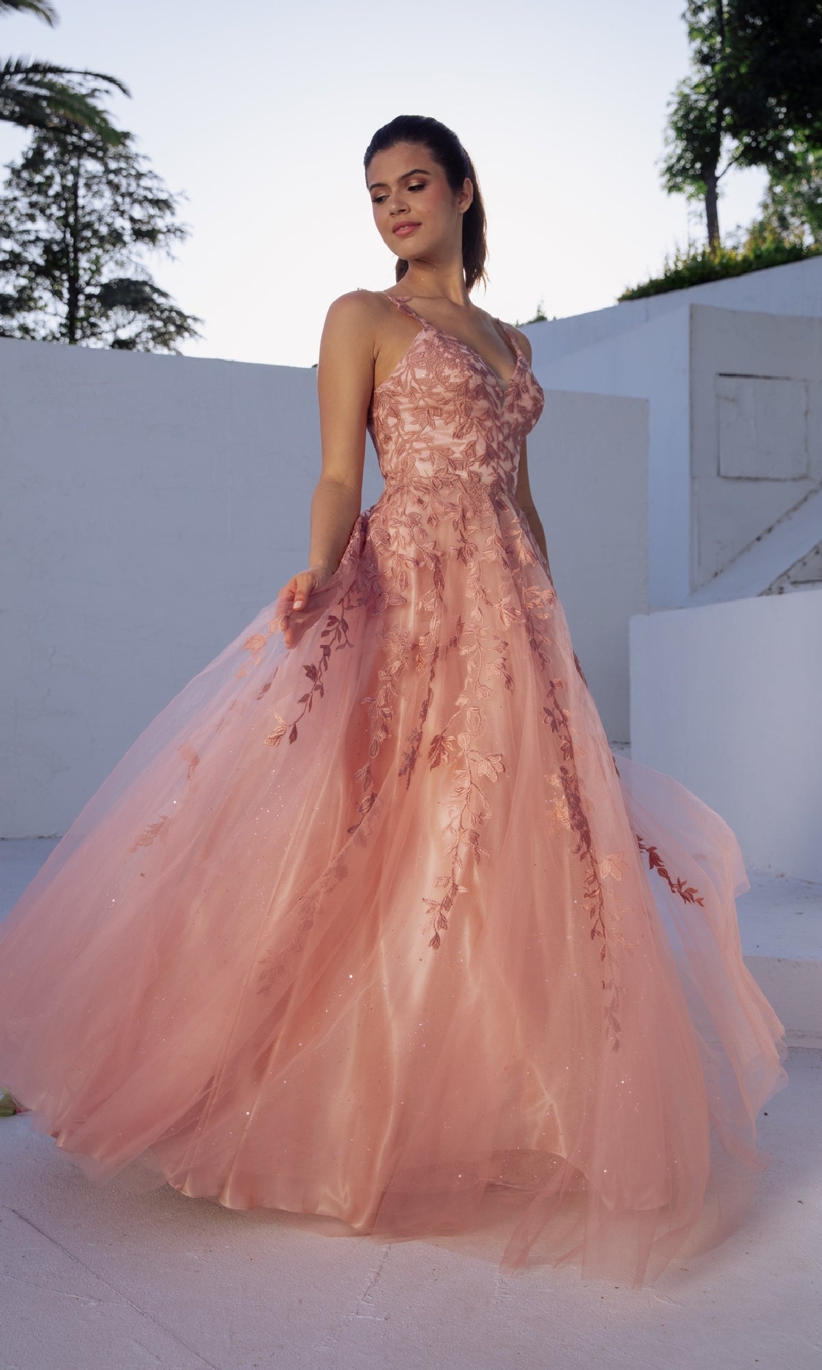 A woman stands outdoors on a white surface, wearing the Embroidered-Lace Long Prom Dress: Eureka 9858—a pink, sleeveless, floor-length formal dress with floral lace details—while looking slightly to the side.
