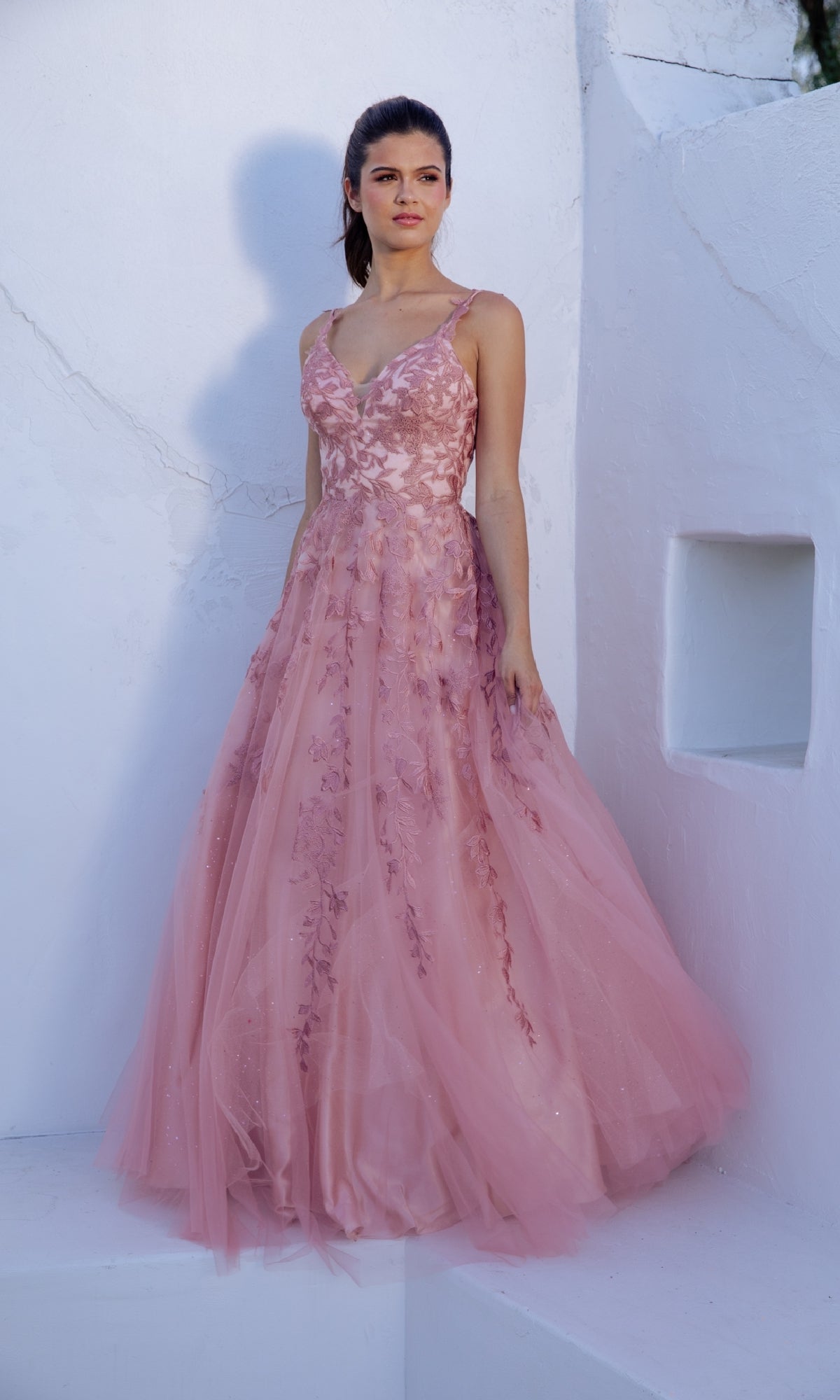 A woman stands against a white wall, wearing the Eureka 9858 Embroidered-Lace Long Prom Dress in pink, featuring a sleeveless A-line design with floral embroidery and a flowing tulle skirt.