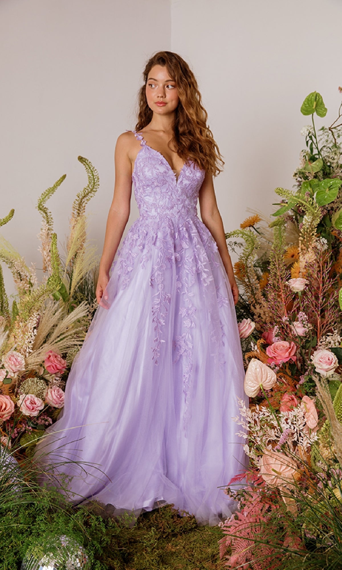 A woman in the Eureka 9858 Embroidered-Lace Long Prom Dress with floral appliqué stands among greenery and pink flowers.
